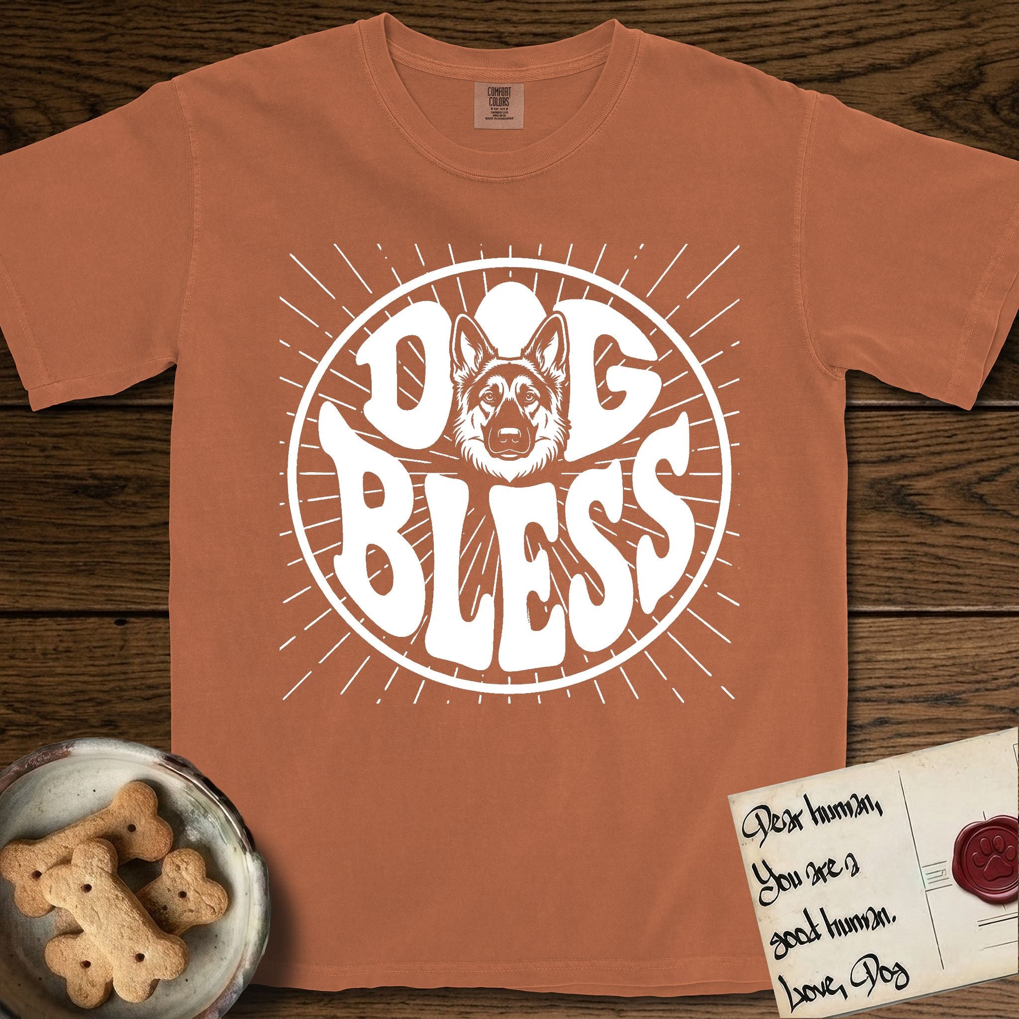 Dog Bless German Shepherd T-Shirt flat lay in Yam, featuring Retro bubble-letter badge reading DOG BLESS with a German Shepherd face with tall upright ears framed in the letter O, hand-drawn sunburst rays radiating outward.