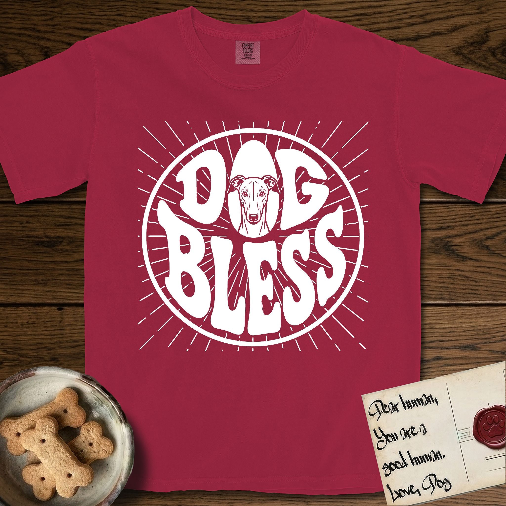Dog Bless Greyhound T-Shirt flat lay in Chili, featuring Retro bubble-letter badge reading DOG BLESS with a slender Greyhound face with folded rose ears framed in the letter O, hand-drawn sunburst rays radiating outward.