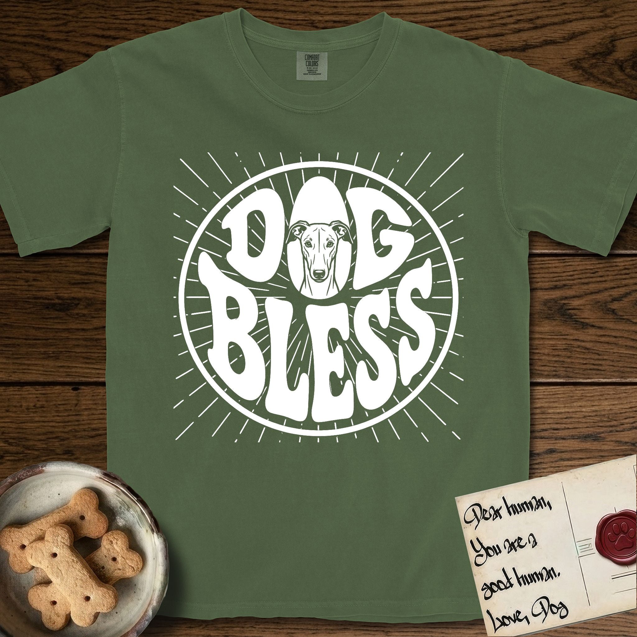 Dog Bless Greyhound T-Shirt flat lay in Moss, featuring Retro bubble-letter badge reading DOG BLESS with a slender Greyhound face with folded rose ears framed in the letter O, hand-drawn sunburst rays radiating outward.