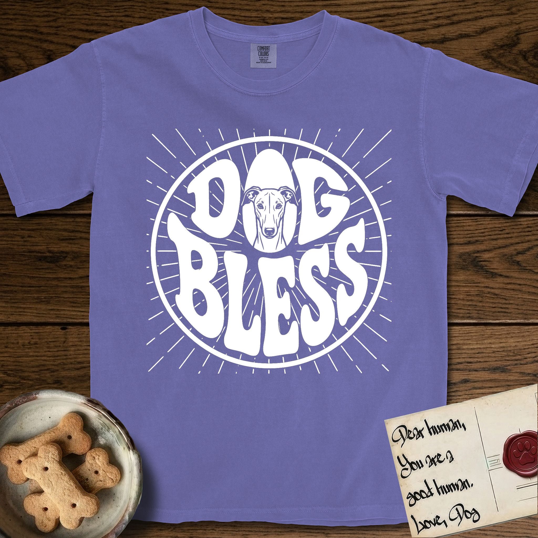 Dog Bless Greyhound T-Shirt flat lay in Violet, featuring Retro bubble-letter badge reading DOG BLESS with a slender Greyhound face with folded rose ears framed in the letter O, hand-drawn sunburst rays radiating outward.