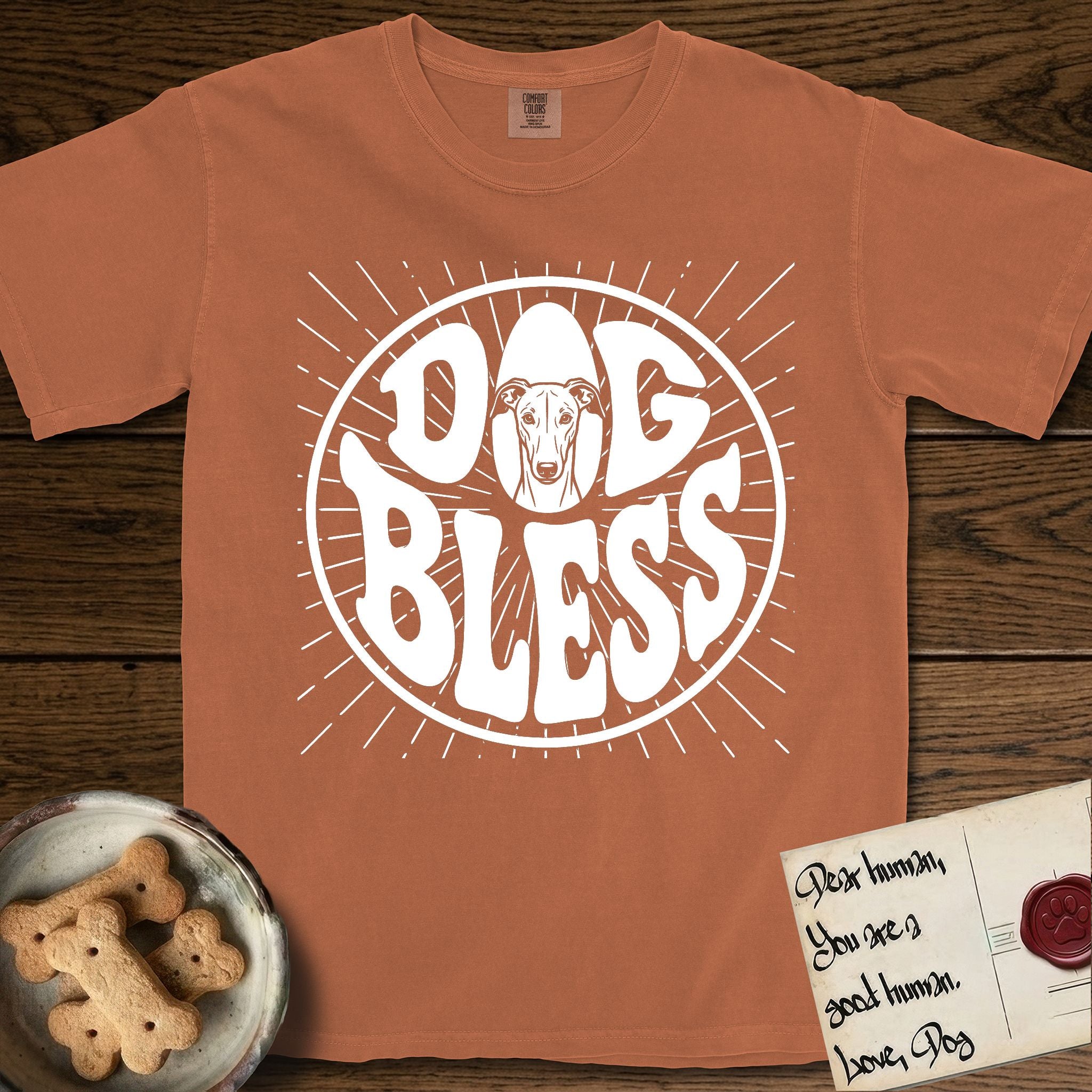 Dog Bless Greyhound T-Shirt flat lay in Yam, featuring Retro bubble-letter badge reading DOG BLESS with a slender Greyhound face with folded rose ears framed in the letter O, hand-drawn sunburst rays radiating outward.