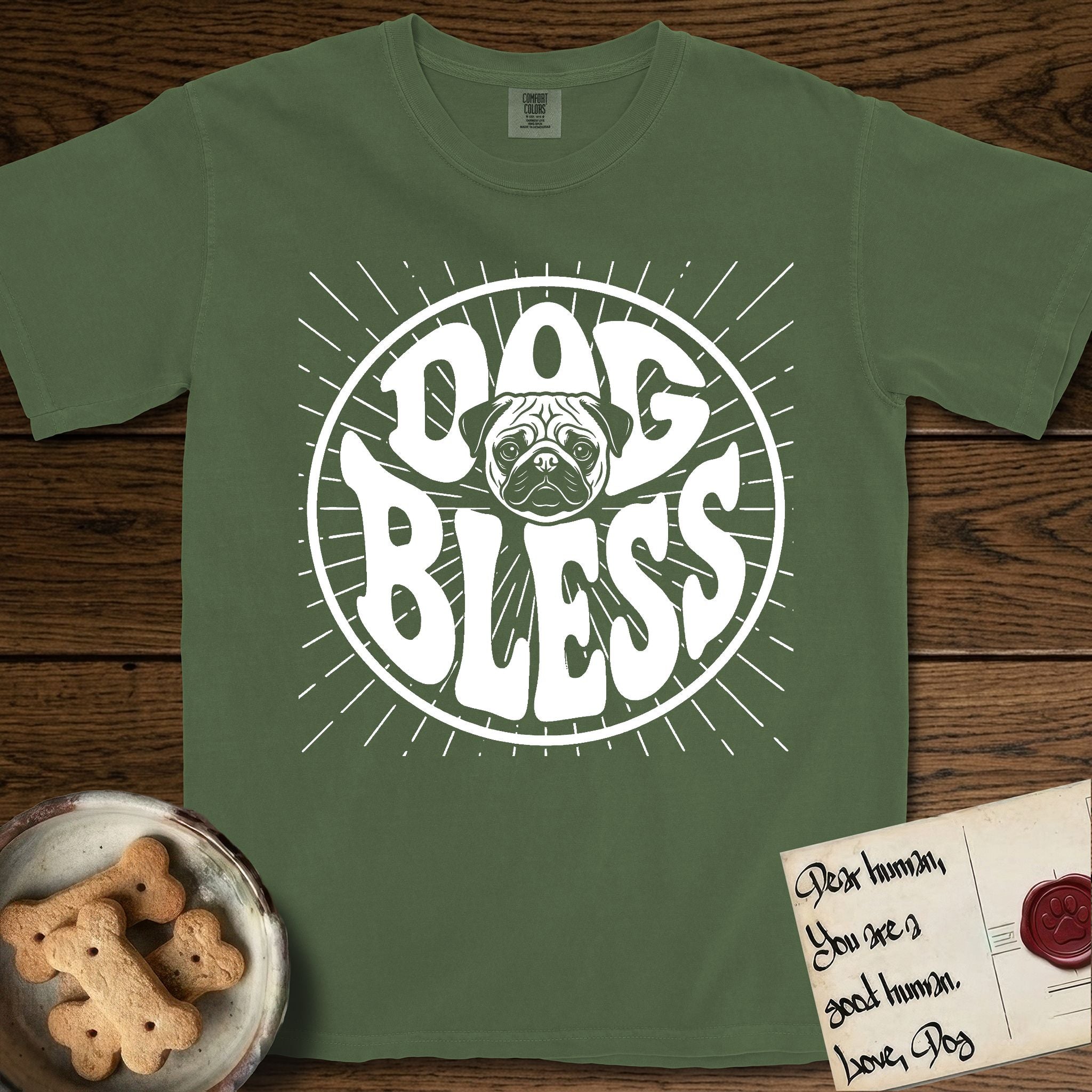 Dog Bless Pug T-Shirt flat lay in Moss, featuring Retro bubble-letter badge reading DOG BLESS with a wrinkled Pug face with large round eyes framed in the letter O, hand-drawn sunburst rays radiating outward.
