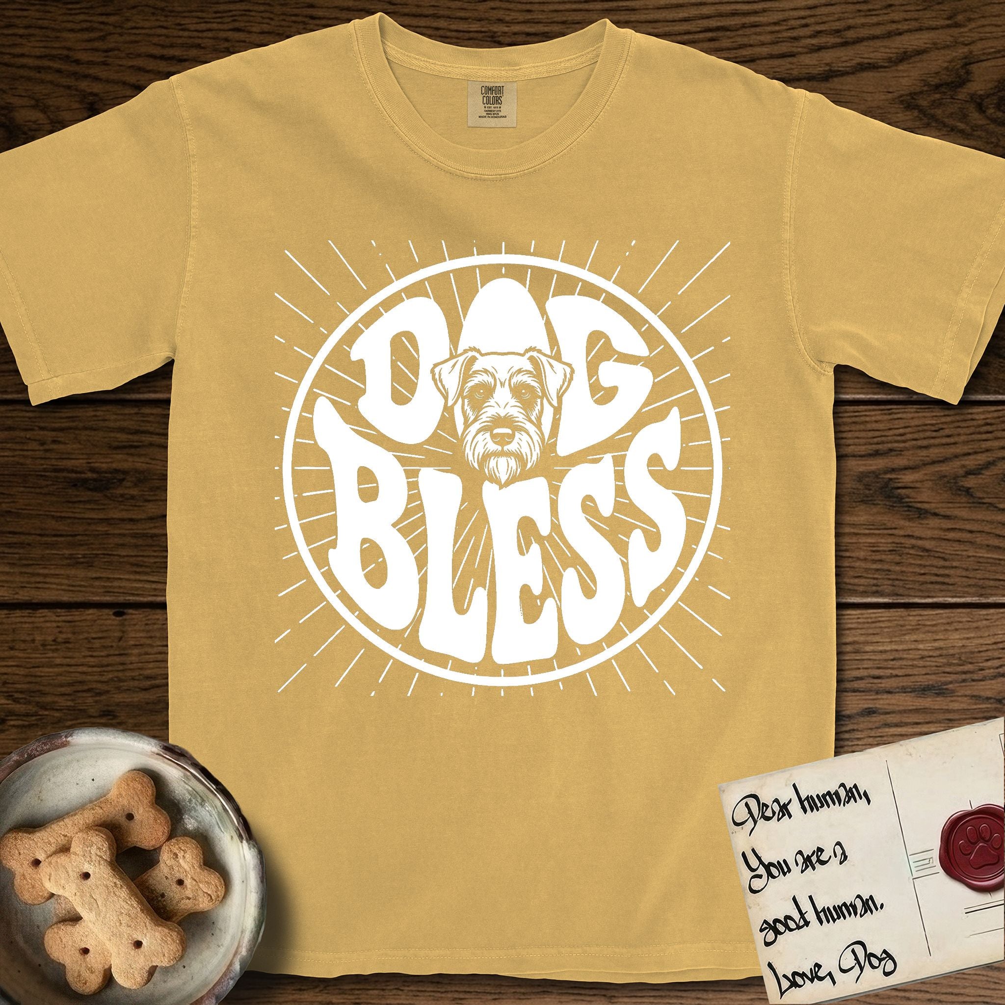 Dog Bless Schnauzer T-Shirt flat lay in Mustard, featuring Retro bubble-letter badge reading DOG BLESS with a bearded Schnauzer face with bushy eyebrows framed in the letter O, hand-drawn sunburst rays radiating outward.
