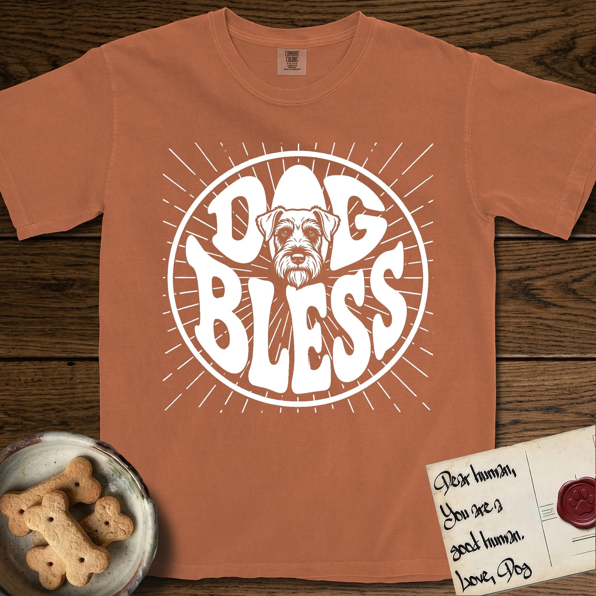 Dog Bless Schnauzer T-Shirt flat lay in Yam, featuring Retro bubble-letter badge reading DOG BLESS with a bearded Schnauzer face with bushy eyebrows framed in the letter O, hand-drawn sunburst rays radiating outward.