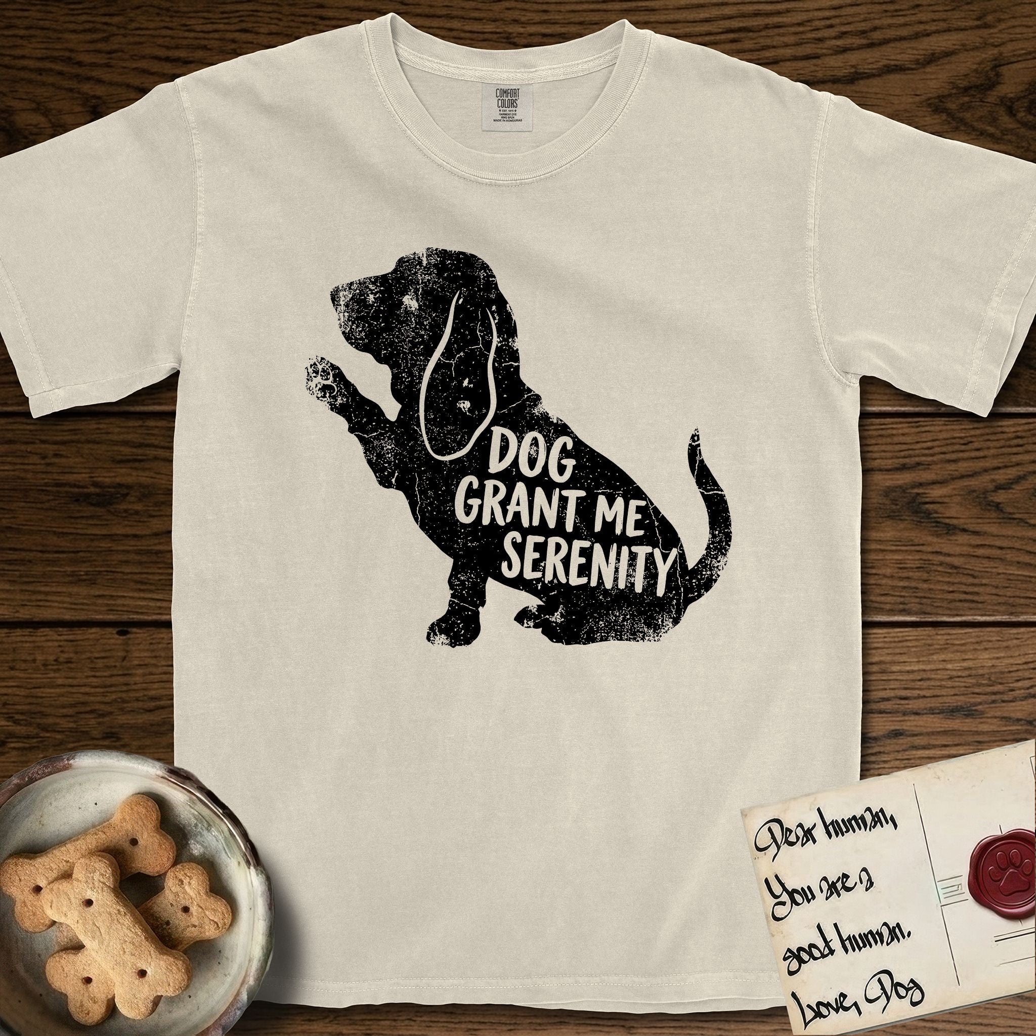 Serenity Basset Hound T-Shirt flat lay in Ivory, featuring Distressed black screen print silhouette of a Basset Hound sitting in profile with one paw raised and long ears hanging, the words DOG GRANT ME SERENITY showing through his body in the shirt color.
