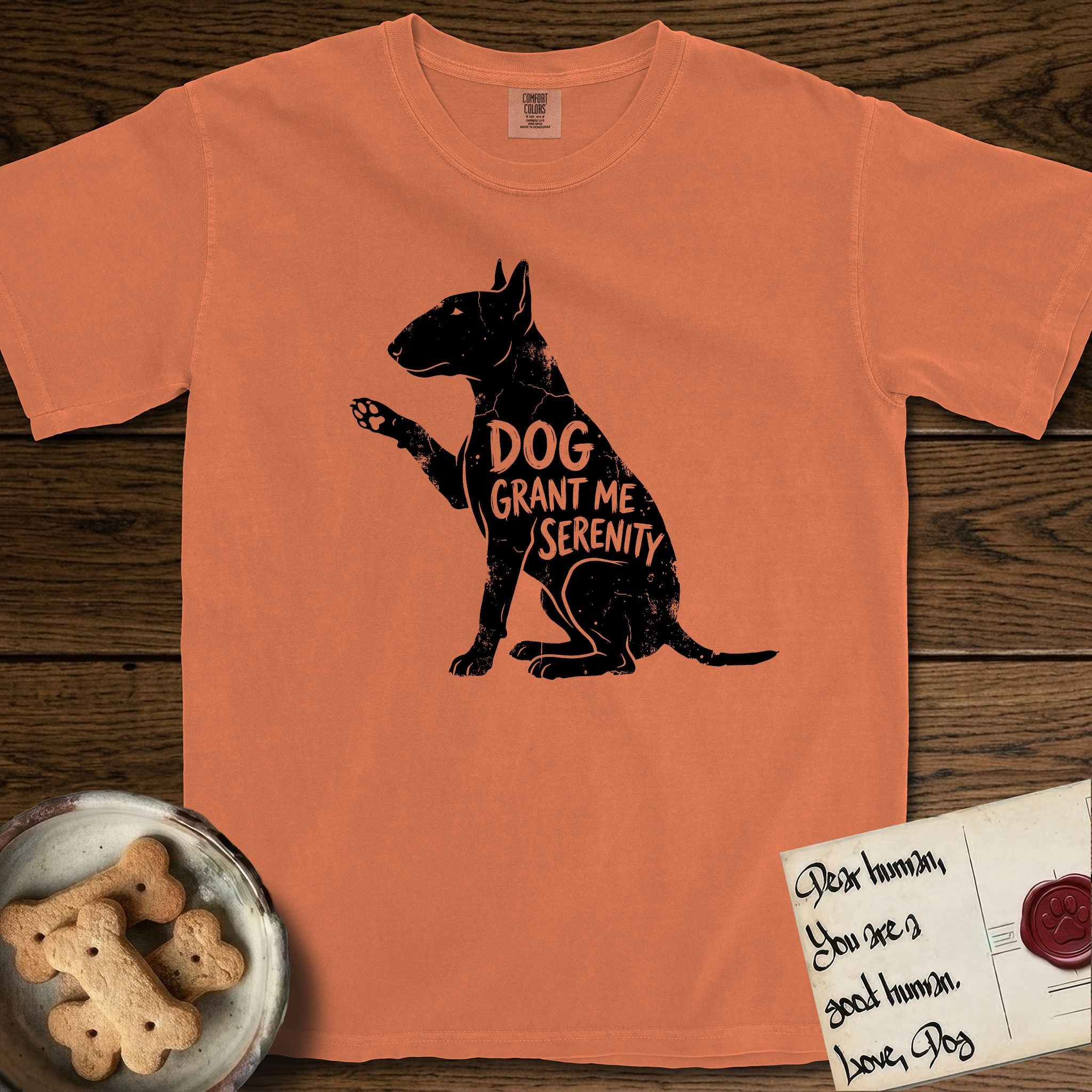 Serenity Bull Terrier T-Shirt flat lay in Melon, featuring Distressed black screen print silhouette of a Bull Terrier sitting in profile with one paw raised and an egg-shaped head, the words DOG GRANT ME SERENITY showing through her body in the shirt color.