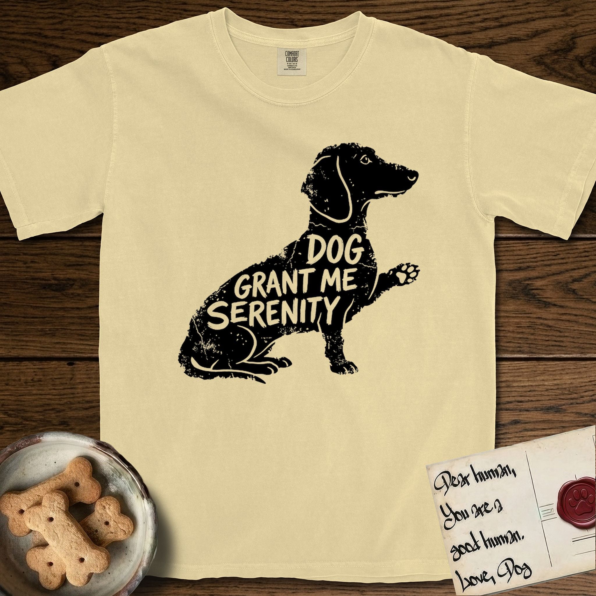 Serenity Dachshund T-Shirt flat lay in Butter, featuring Distressed black screen print silhouette of a Dachshund sitting in profile with one paw raised, a long low body and long hanging ears, the words DOG GRANT ME SERENITY showing through her body in the shirt color.