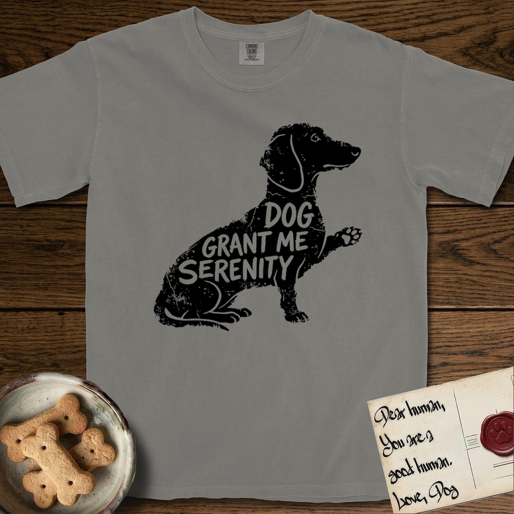 Serenity Dachshund T-Shirt flat lay in Grey, featuring Distressed black screen print silhouette of a Dachshund sitting in profile with one paw raised, a long low body and long hanging ears, the words DOG GRANT ME SERENITY showing through her body in the shirt color.