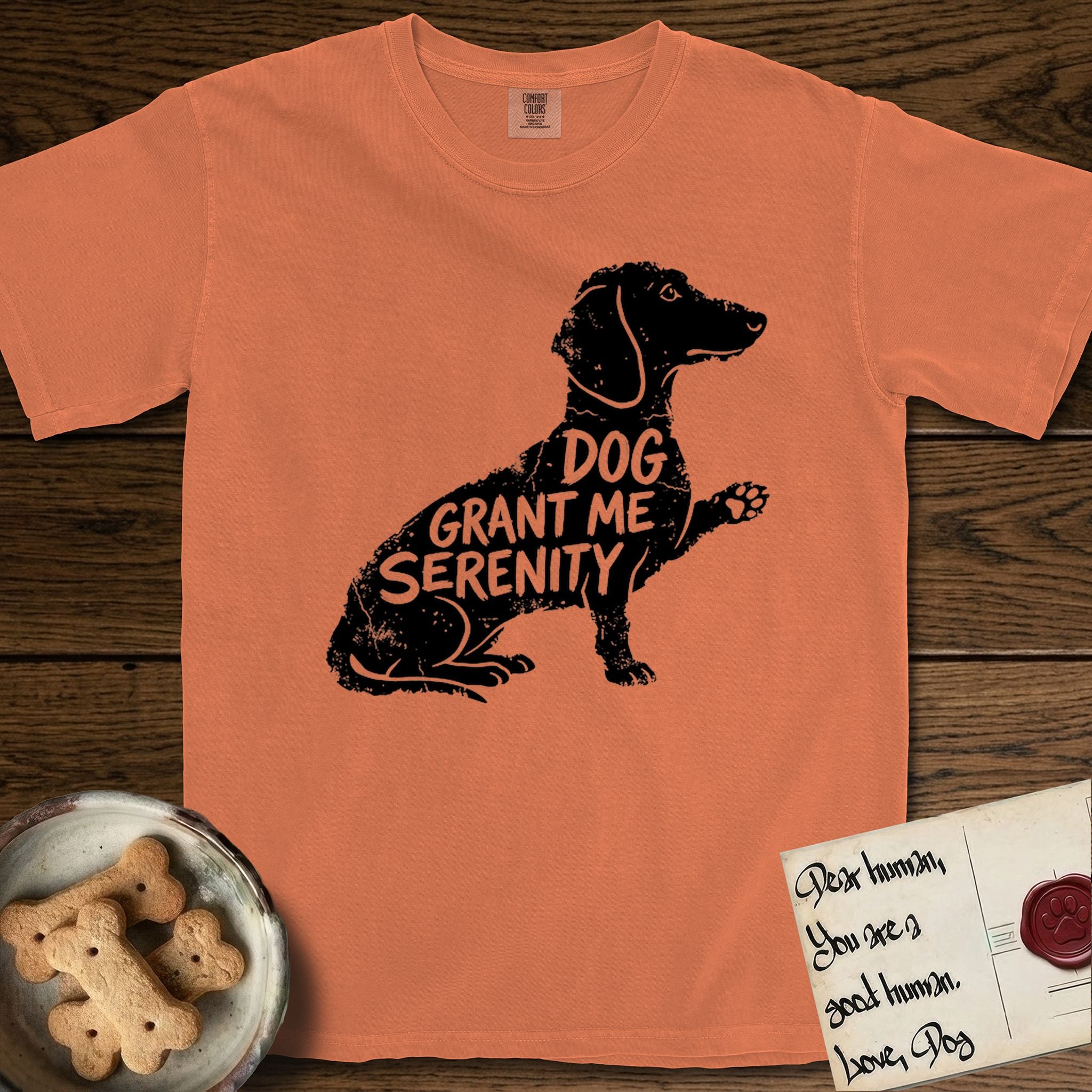 Serenity Dachshund T-Shirt flat lay in Melon, featuring Distressed black screen print silhouette of a Dachshund sitting in profile with one paw raised, a long low body and long hanging ears, the words DOG GRANT ME SERENITY showing through her body in the shirt color.