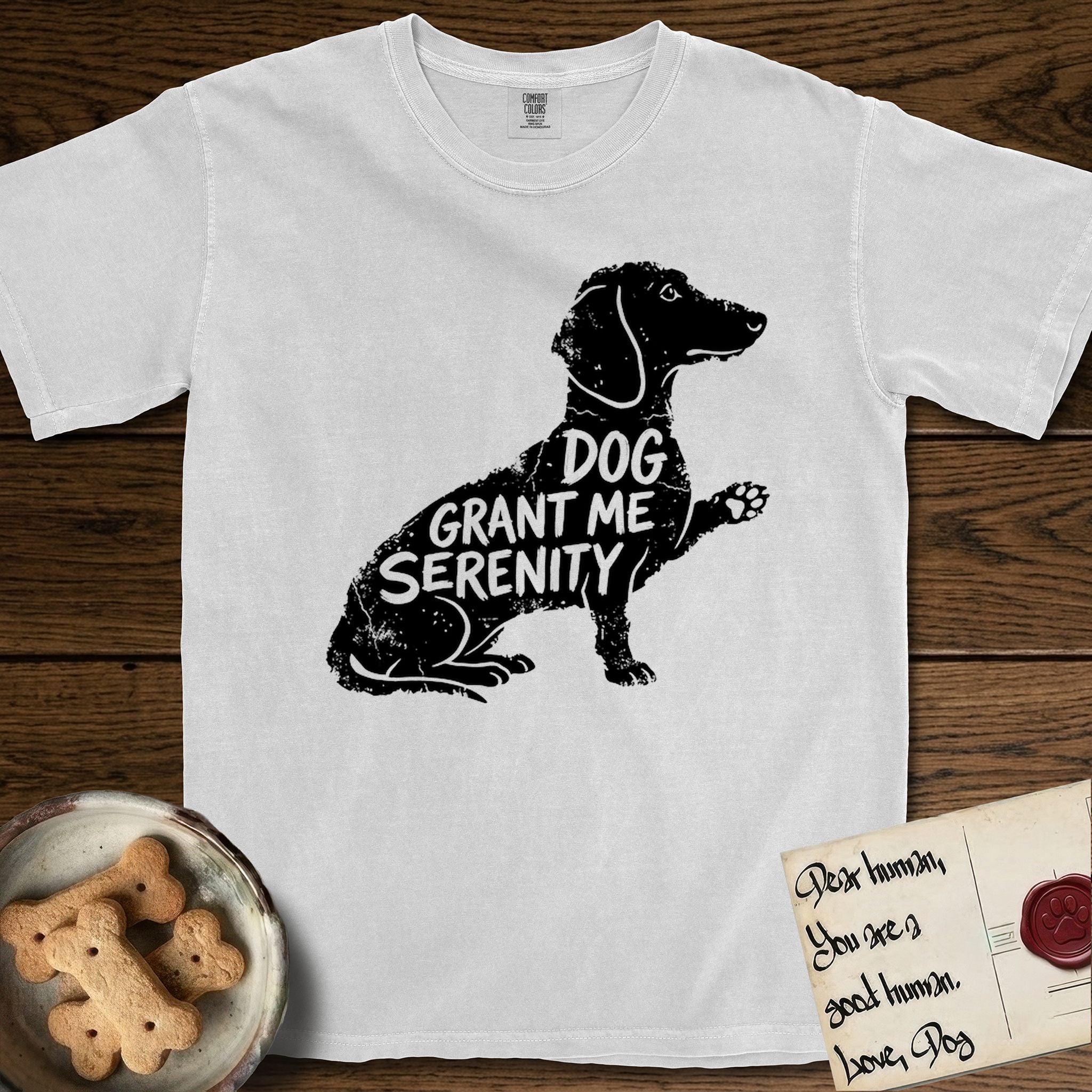 Serenity Dachshund T-Shirt flat lay in White, featuring Distressed black screen print silhouette of a Dachshund sitting in profile with one paw raised, a long low body and long hanging ears, the words DOG GRANT ME SERENITY showing through her body in the shirt color.