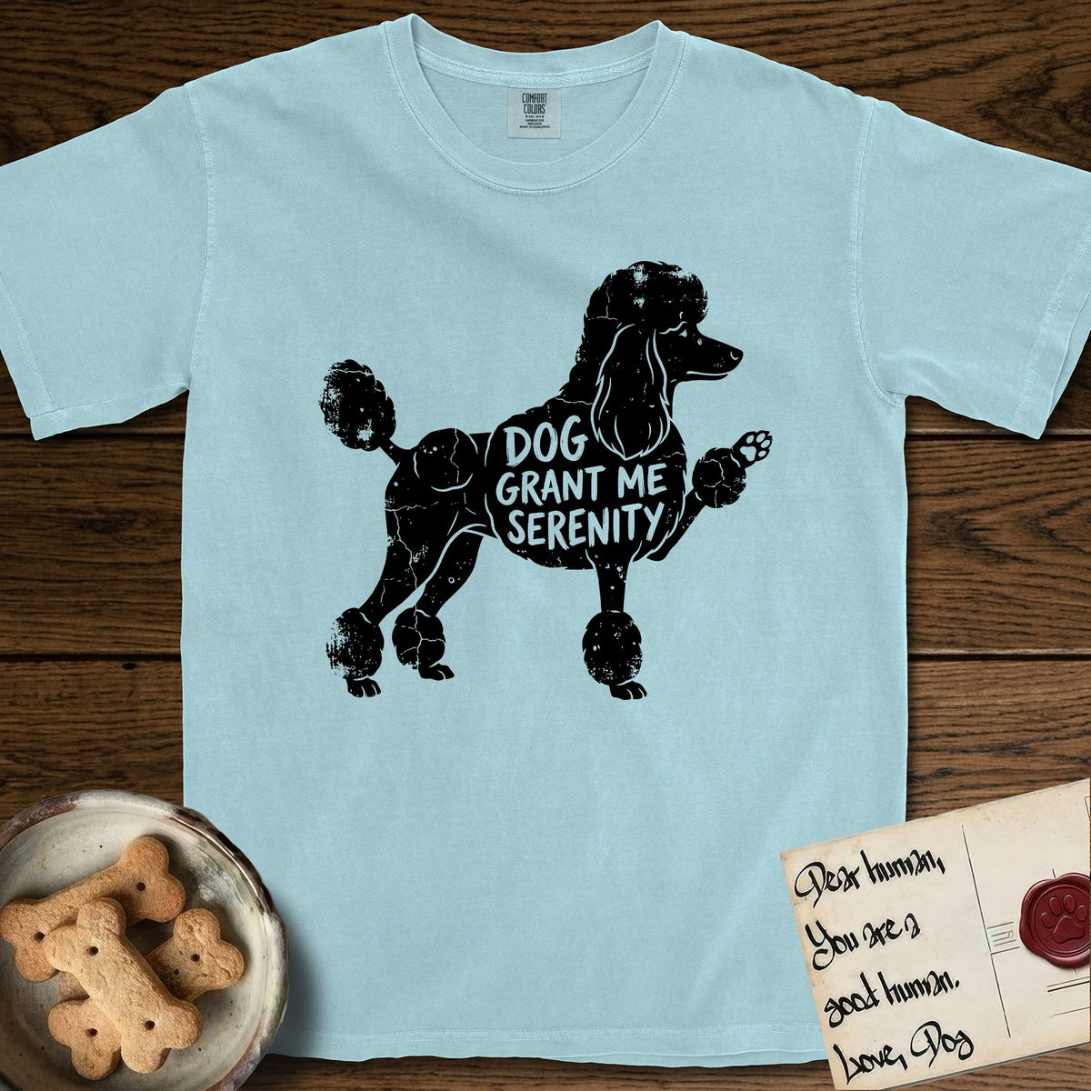 Serenity Poodle T-Shirt flat lay in Chambray, featuring Distressed black screen print silhouette of a Poodle standing in profile with one paw raised, in a continental clip with a topknot and pompons, the words DOG GRANT ME SERENITY showing through her body in the shirt color.