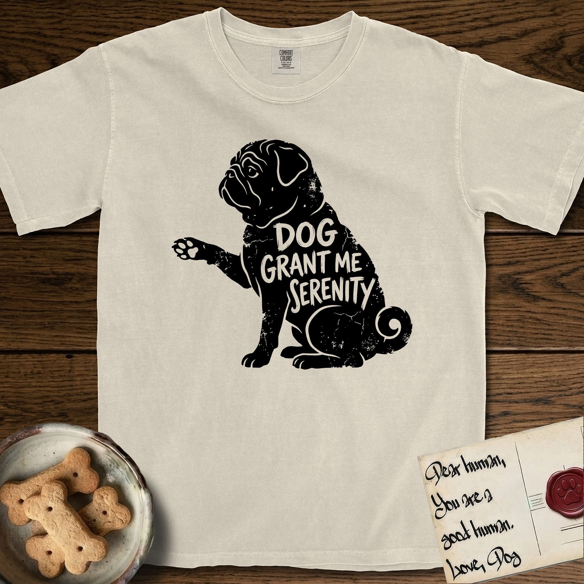 Serenity Pug T-Shirt flat lay in Ivory, featuring Distressed black screen print silhouette of a Pug sitting in profile with one paw raised, a wrinkled face and a tightly curled tail, the words DOG GRANT ME SERENITY showing through his body in the shirt color.