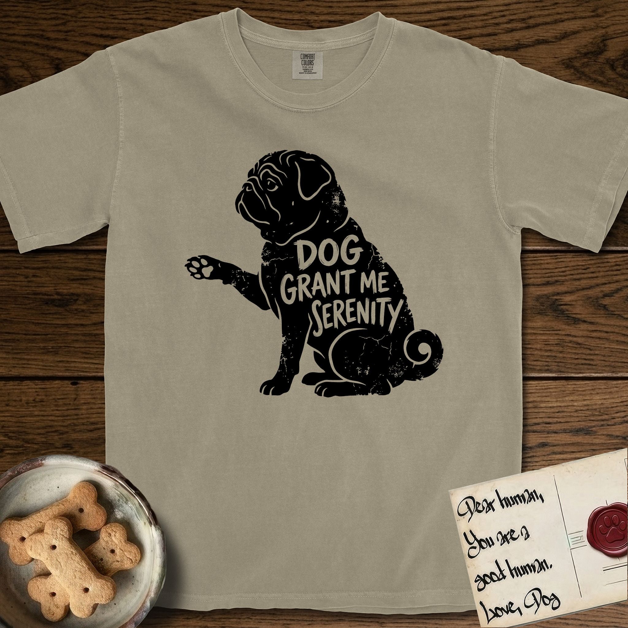 Serenity Pug T-Shirt flat lay in Khaki, featuring Distressed black screen print silhouette of a Pug sitting in profile with one paw raised, a wrinkled face and a tightly curled tail, the words DOG GRANT ME SERENITY showing through his body in the shirt color.