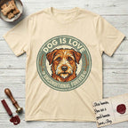 Dog is Love T-Shirt flat lay in Natural, featuring a vintage distressed circular badge with a scruffy terrier on a sage-green ground, the words Dog Is Love and Unconditional Forever, and flanking paw prints.