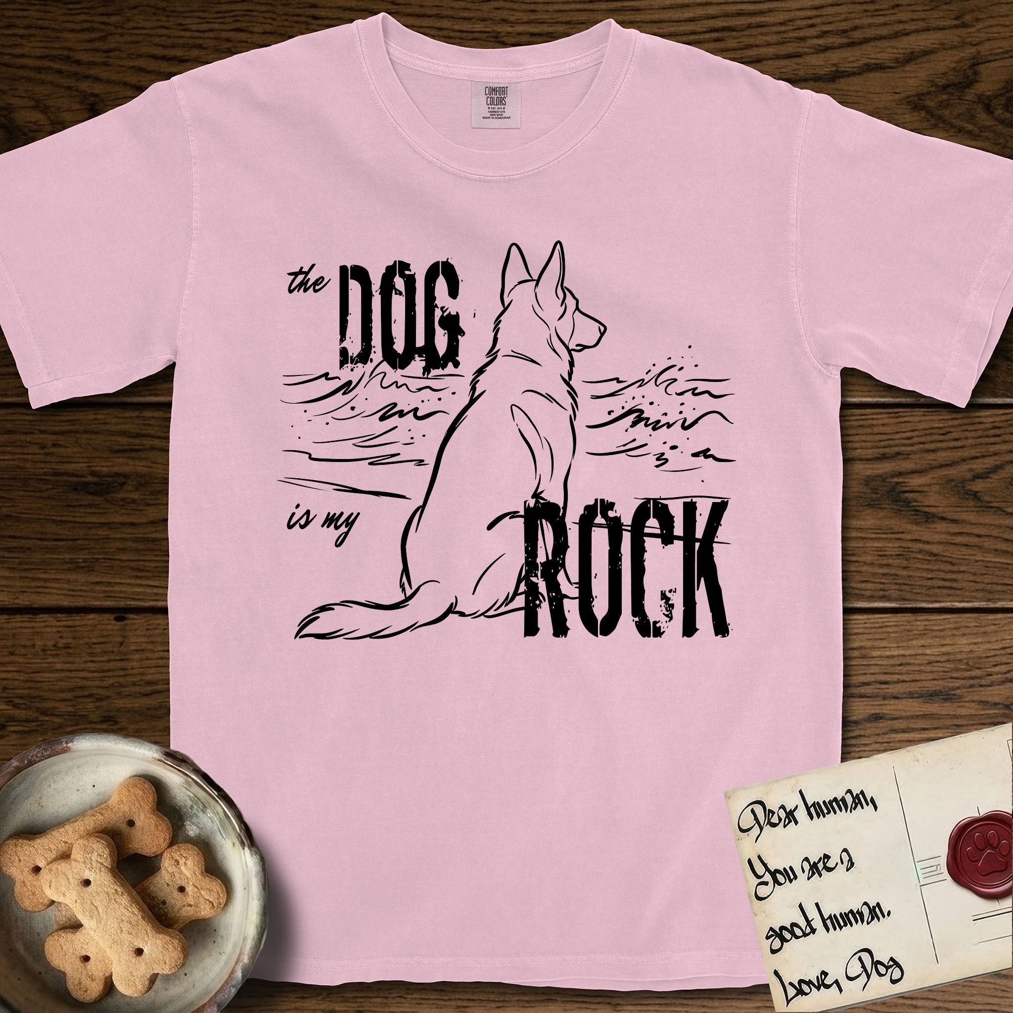 DIMR German Shepherd T-Shirt flat lay in Blossom, featuring ink-line illustration of a German Shepherd seen from behind, sitting at the waterline and watching a stormy sea, with the words the Dog is my Rock.