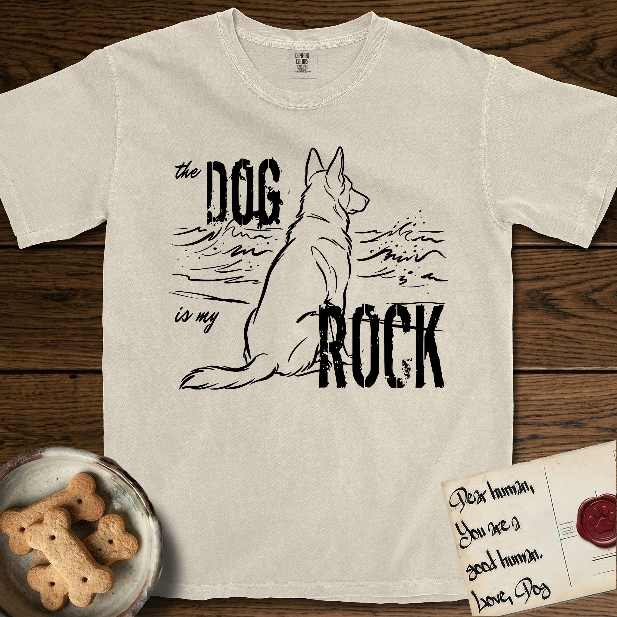 DIMR German Shepherd T-Shirt flat lay in Ivory, featuring ink-line illustration of a German Shepherd seen from behind, sitting at the waterline and watching a stormy sea, with the words the Dog is my Rock.
