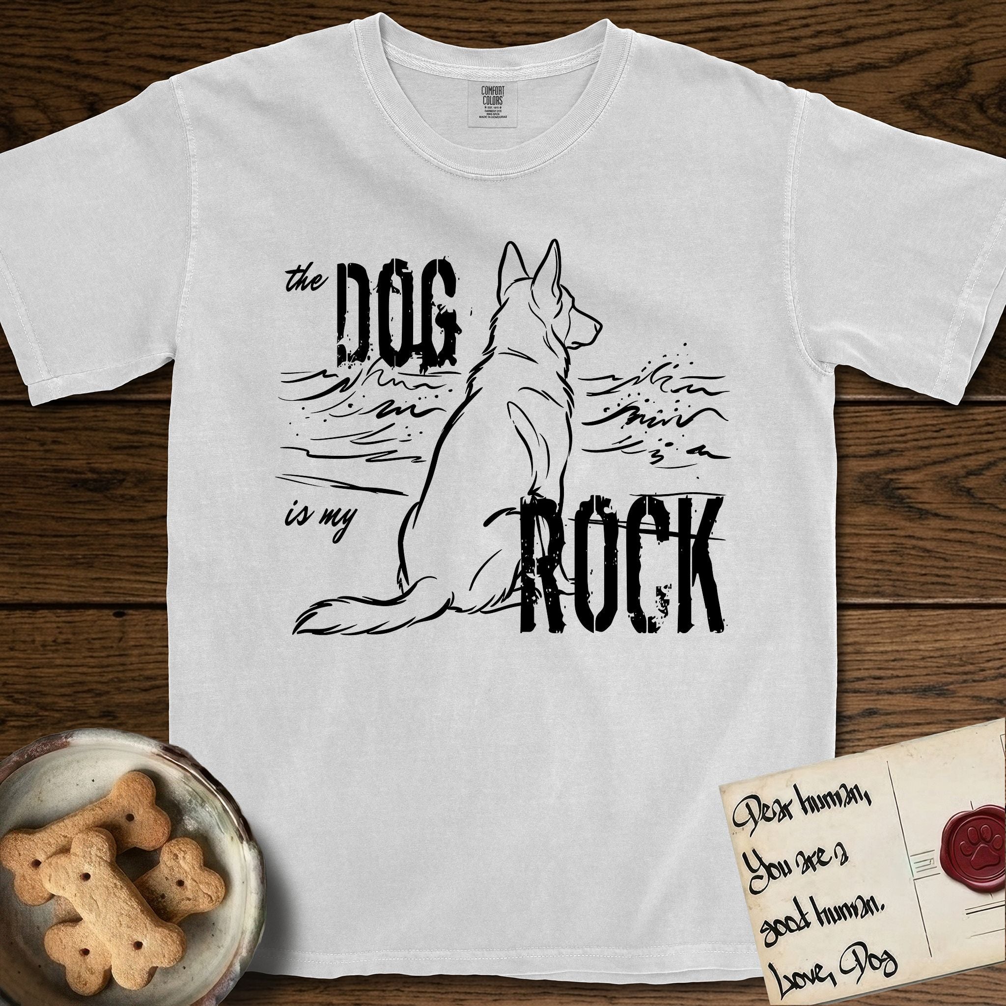DIMR German Shepherd T-Shirt flat lay in White, featuring ink-line illustration of a German Shepherd seen from behind, sitting at the waterline and watching a stormy sea, with the words the Dog is my Rock.