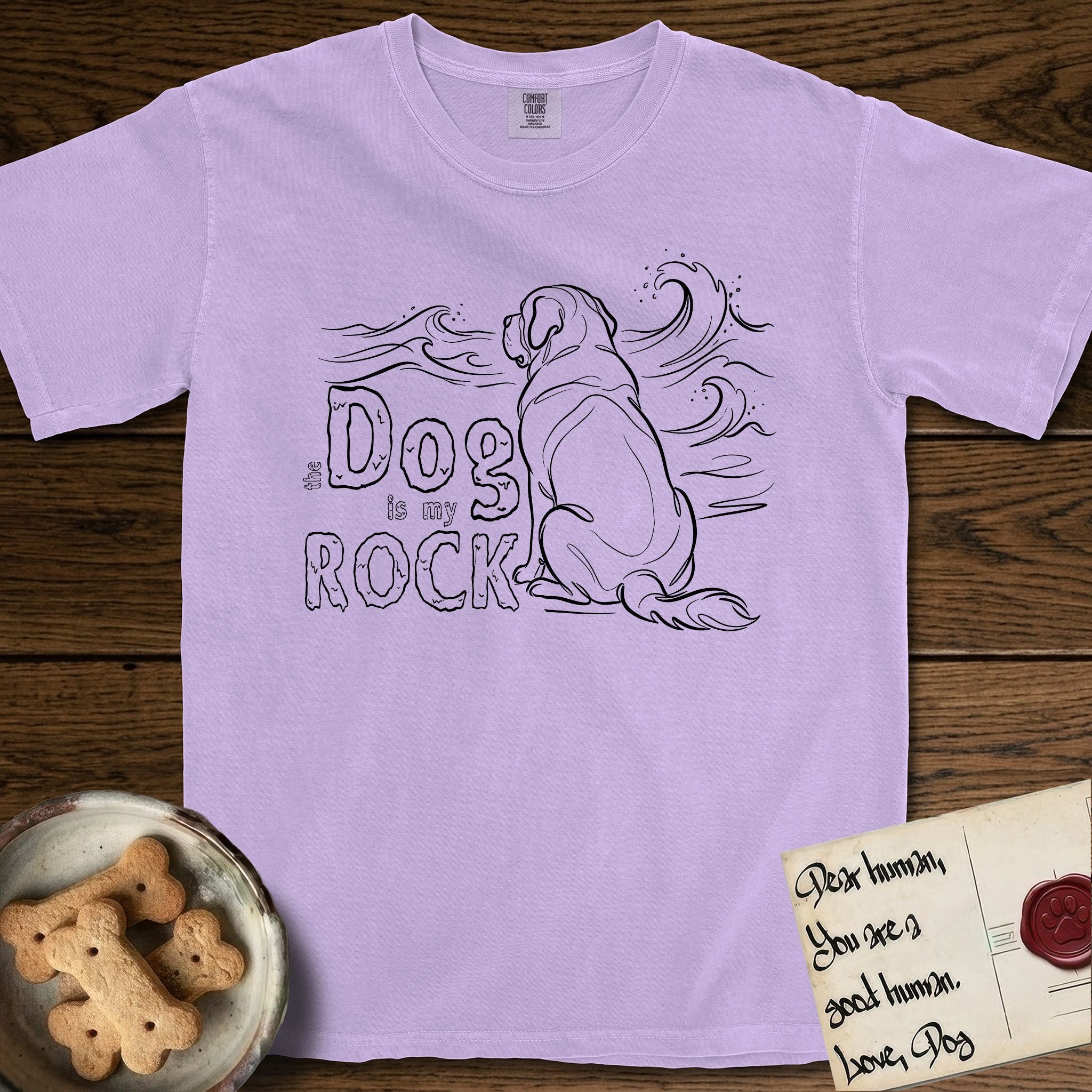 DIMR Saint Bernard T-Shirt flat lay in Orchid, featuring ink-line illustration of a Saint Bernard seen from behind, sitting at the water's edge and watching a stormy sea, with the words the Dog is my Rock.