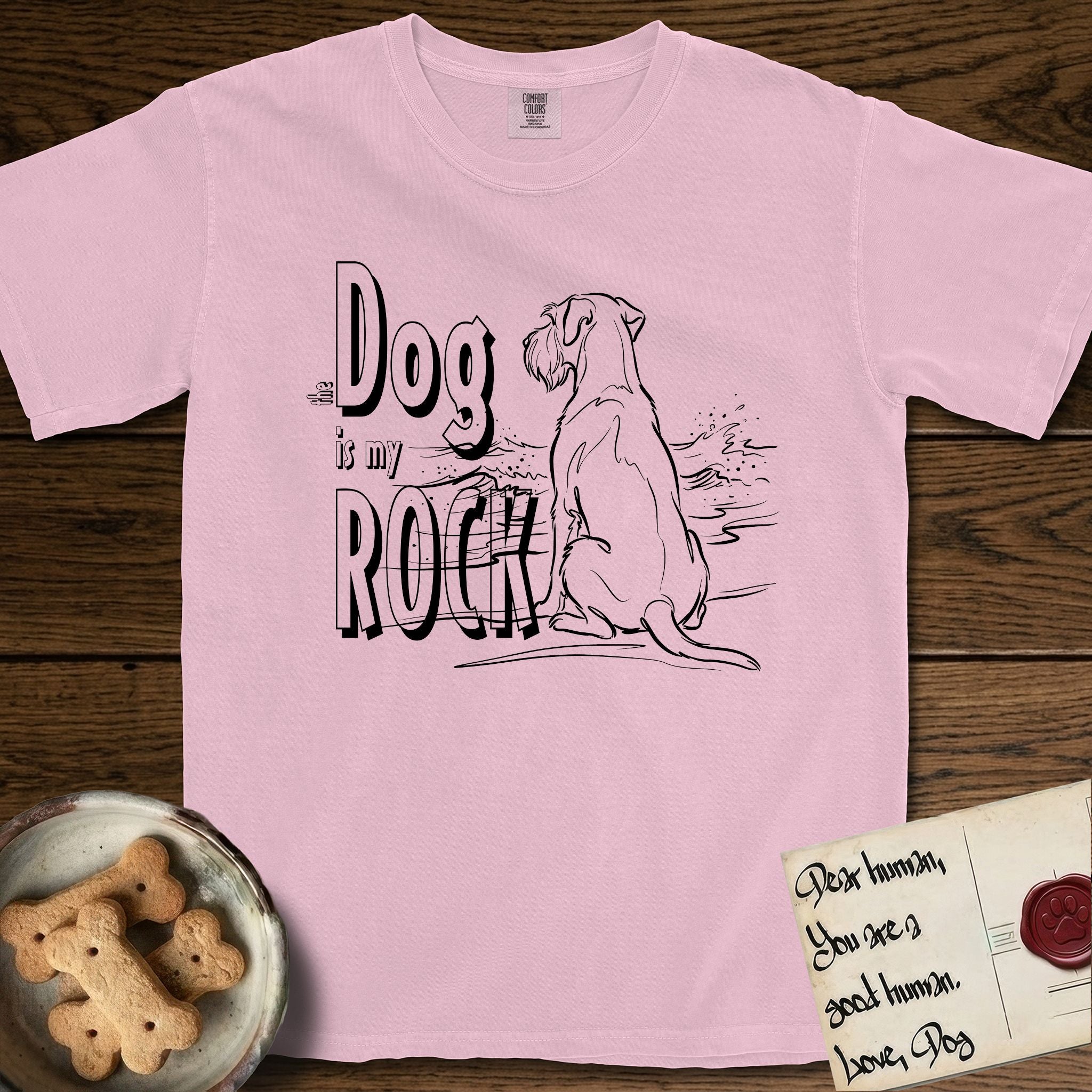 DIMR Schnauzer T-Shirt flat lay in Blossom, featuring ink-line illustration of a Schnauzer seen from behind at the water's edge, head turned toward a stormy sea, with the words the Dog is my Rock.