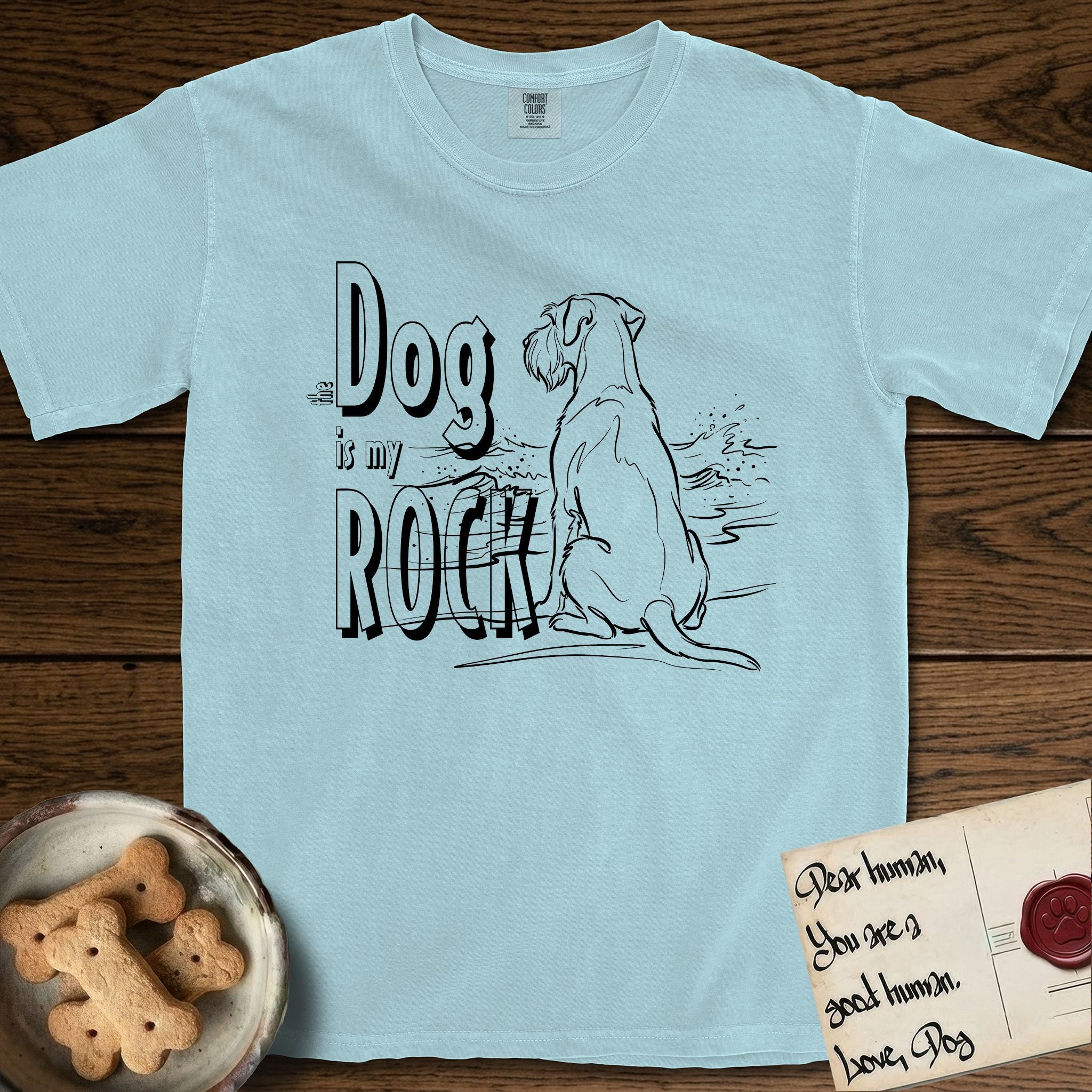 DIMR Schnauzer T-Shirt flat lay in Chambray, featuring ink-line illustration of a Schnauzer seen from behind at the water's edge, head turned toward a stormy sea, with the words the Dog is my Rock.