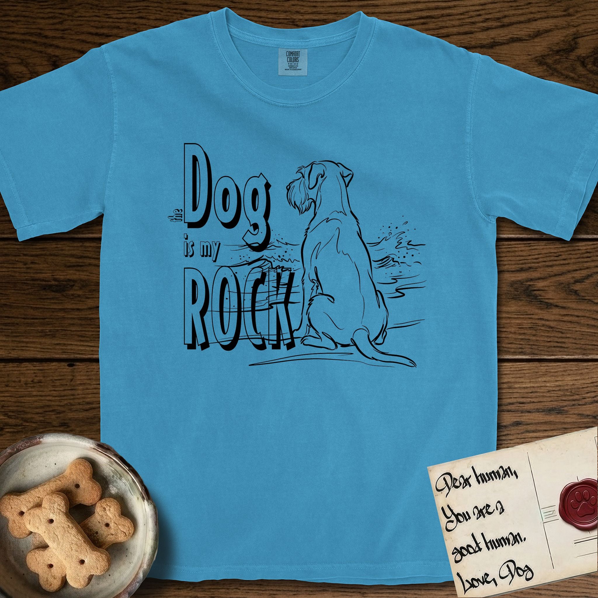 DIMR Schnauzer T-Shirt flat lay in Royal Caribe, featuring ink-line illustration of a Schnauzer seen from behind at the water's edge, head turned toward a stormy sea, with the words the Dog is my Rock.