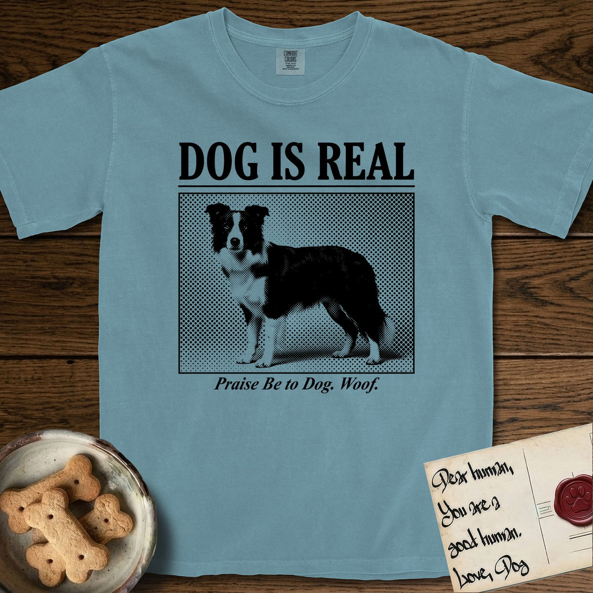 Dog is Real Border Collie T-Shirt flat lay in Ice Blue, featuring Black halftone newspaper photo of a black and white Border Collie standing in profile with head turned to the camera, under the headline DOG IS REAL, with the caption Praise Be to Dog. Woof..