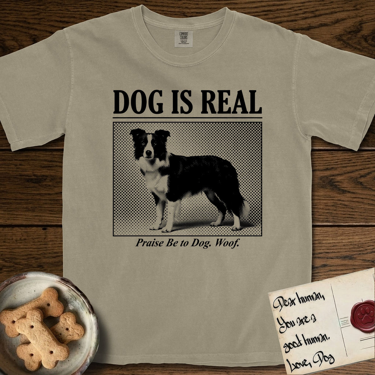 Dog is Real Border Collie T-Shirt flat lay in Khaki, featuring Black halftone newspaper photo of a black and white Border Collie standing in profile with head turned to the camera, under the headline DOG IS REAL, with the caption Praise Be to Dog. Woof..