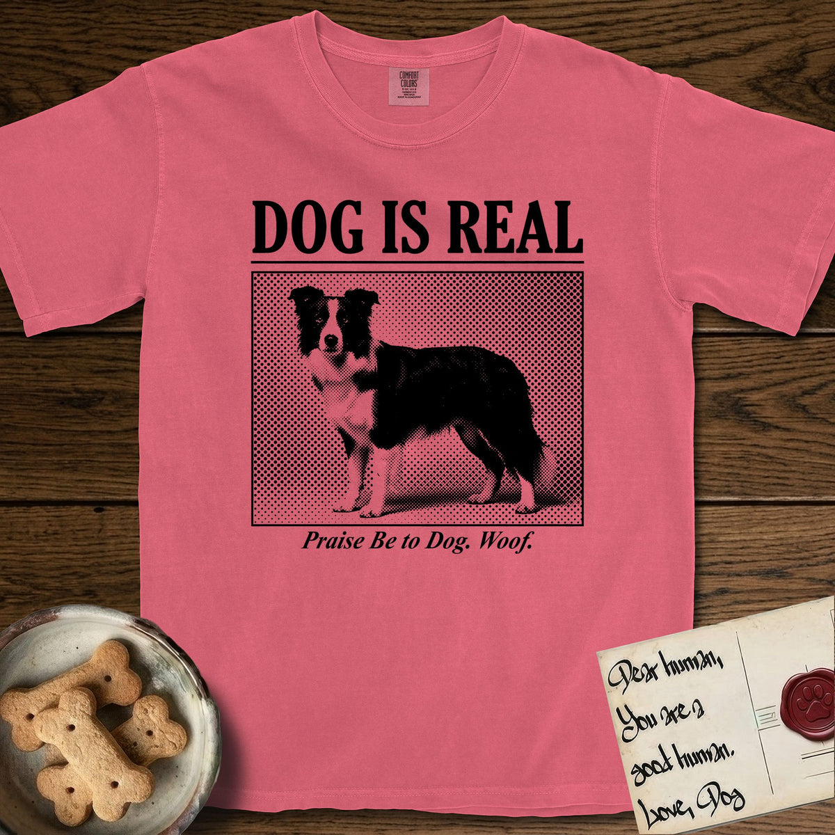 Dog is Real Border Collie T-Shirt flat lay in Watermelon, featuring Black halftone newspaper photo of a black and white Border Collie standing in profile with head turned to the camera, under the headline DOG IS REAL, with the caption Praise Be to Dog. Woof..