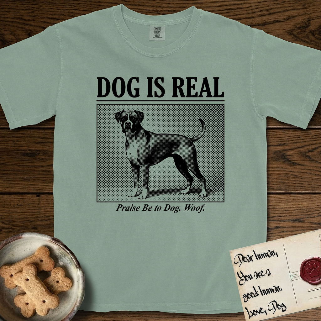 Dog is Real Boxer T-Shirt flat lay in Bay, featuring Black halftone newspaper photo of a Boxer with a white chest standing in profile with head turned to the camera, under the headline DOG IS REAL, with the caption Praise Be to Dog. Woof..