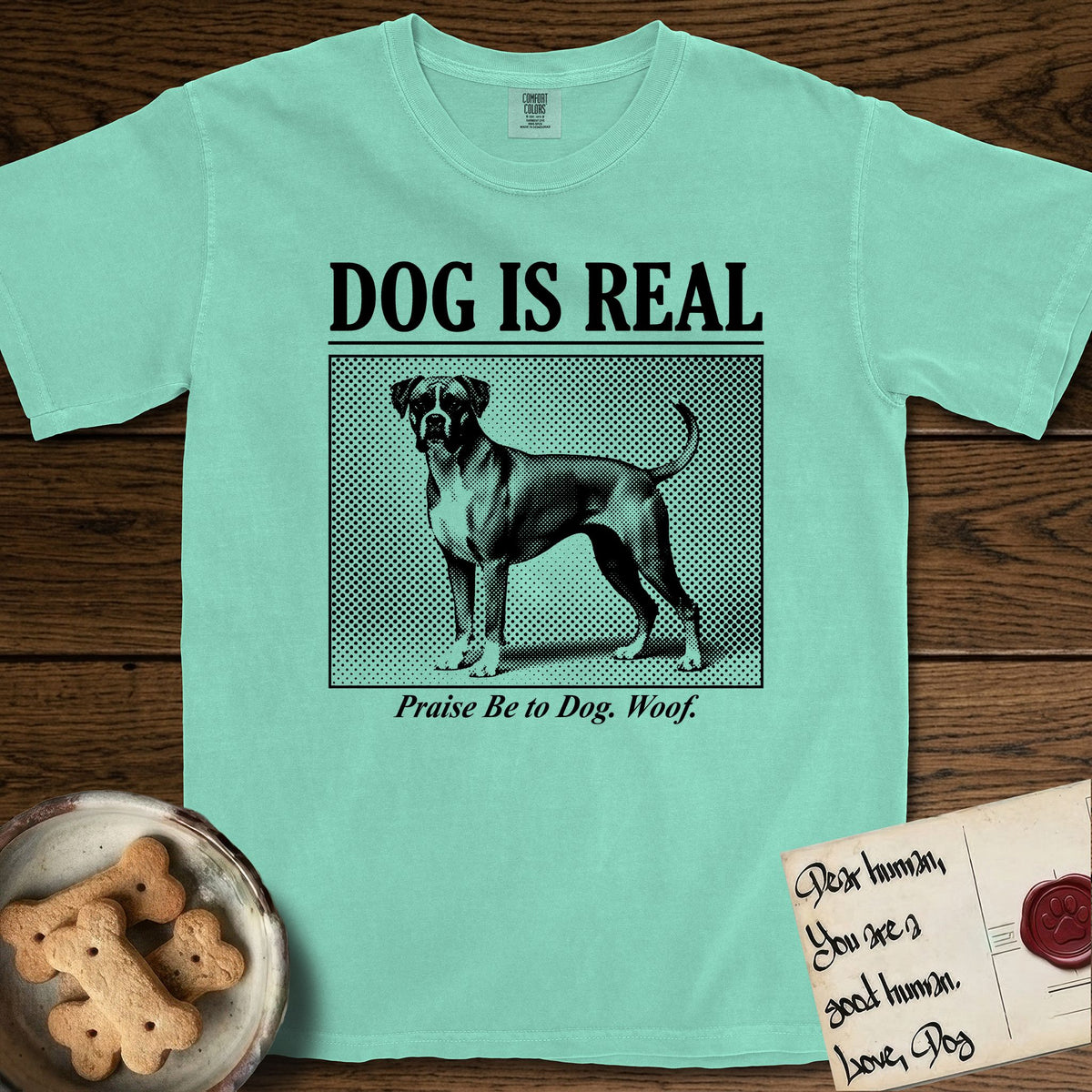 Dog is Real Boxer T-Shirt flat lay in Island Reef, featuring Black halftone newspaper photo of a Boxer with a white chest standing in profile with head turned to the camera, under the headline DOG IS REAL, with the caption Praise Be to Dog. Woof..