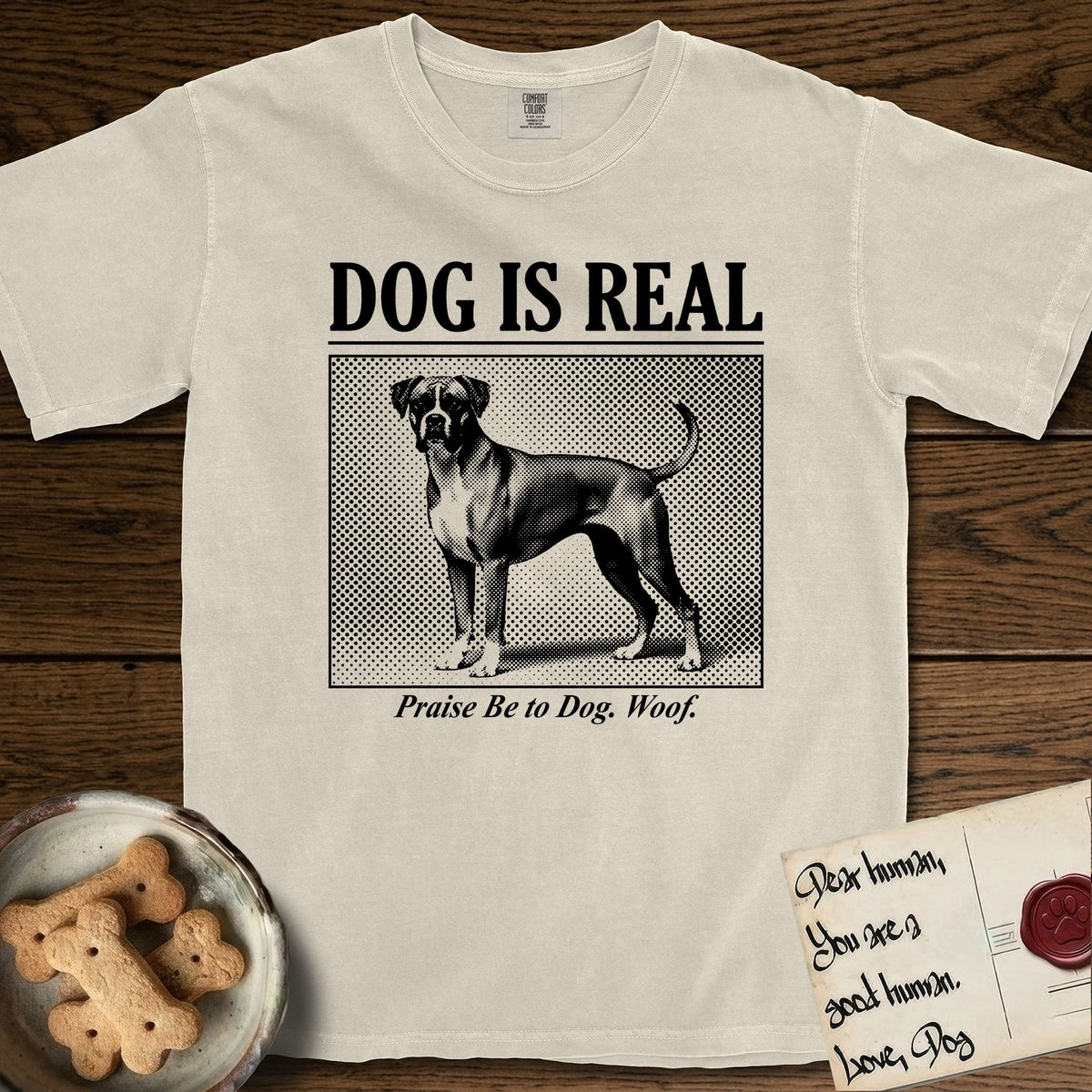 Dog is Real Boxer T-Shirt flat lay in Ivory, featuring Black halftone newspaper photo of a Boxer with a white chest standing in profile with head turned to the camera, under the headline DOG IS REAL, with the caption Praise Be to Dog. Woof..
