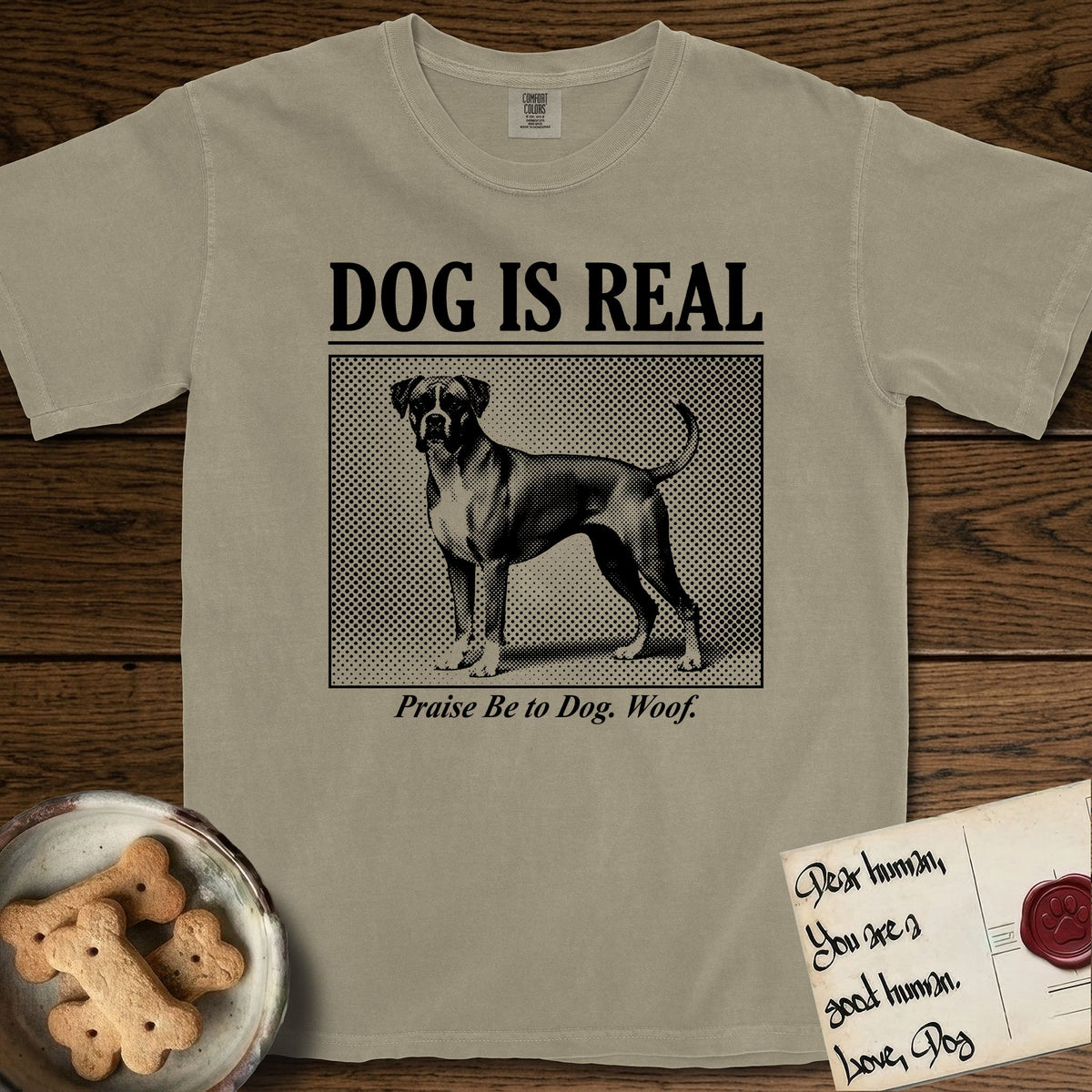 Dog is Real Boxer T-Shirt flat lay in Khaki, featuring Black halftone newspaper photo of a Boxer with a white chest standing in profile with head turned to the camera, under the headline DOG IS REAL, with the caption Praise Be to Dog. Woof..