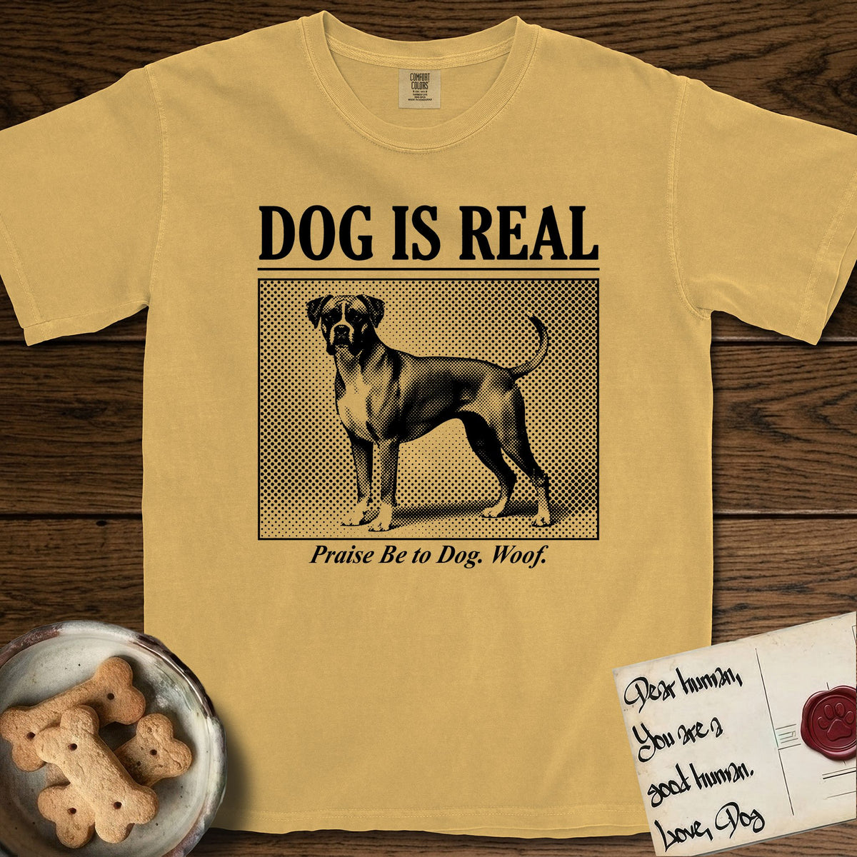 Dog is Real Boxer T-Shirt flat lay in Mustard, featuring Black halftone newspaper photo of a Boxer with a white chest standing in profile with head turned to the camera, under the headline DOG IS REAL, with the caption Praise Be to Dog. Woof..