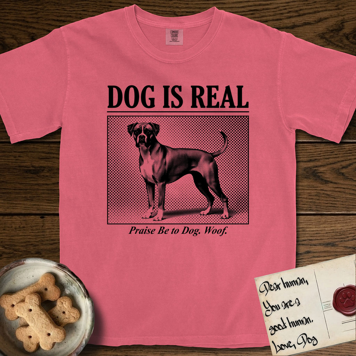 Dog is Real Boxer T-Shirt flat lay in Watermelon, featuring Black halftone newspaper photo of a Boxer with a white chest standing in profile with head turned to the camera, under the headline DOG IS REAL, with the caption Praise Be to Dog. Woof..