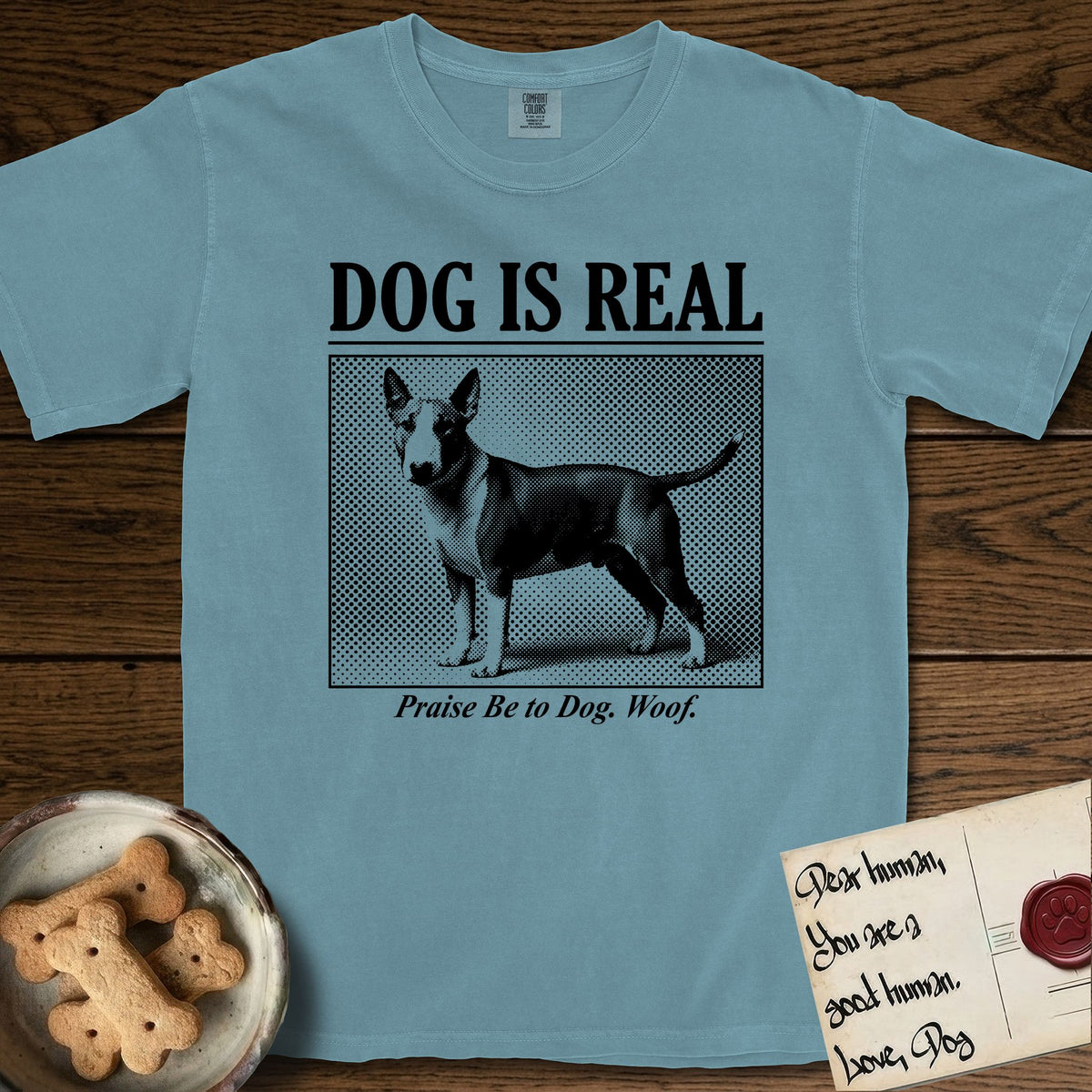 Dog is Real Bull Terrier T-Shirt flat lay in Ice Blue, featuring Black halftone newspaper photo of a Bull Terrier with a white face standing in profile with head turned to the camera, under the headline DOG IS REAL, with the caption Praise Be to Dog. Woof..
