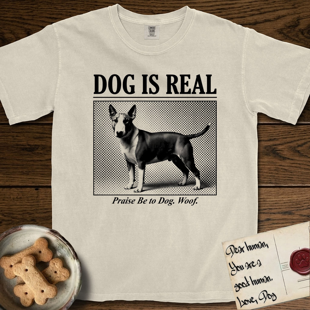 Dog is Real Bull Terrier T-Shirt flat lay in Ivory, featuring Black halftone newspaper photo of a Bull Terrier with a white face standing in profile with head turned to the camera, under the headline DOG IS REAL, with the caption Praise Be to Dog. Woof..