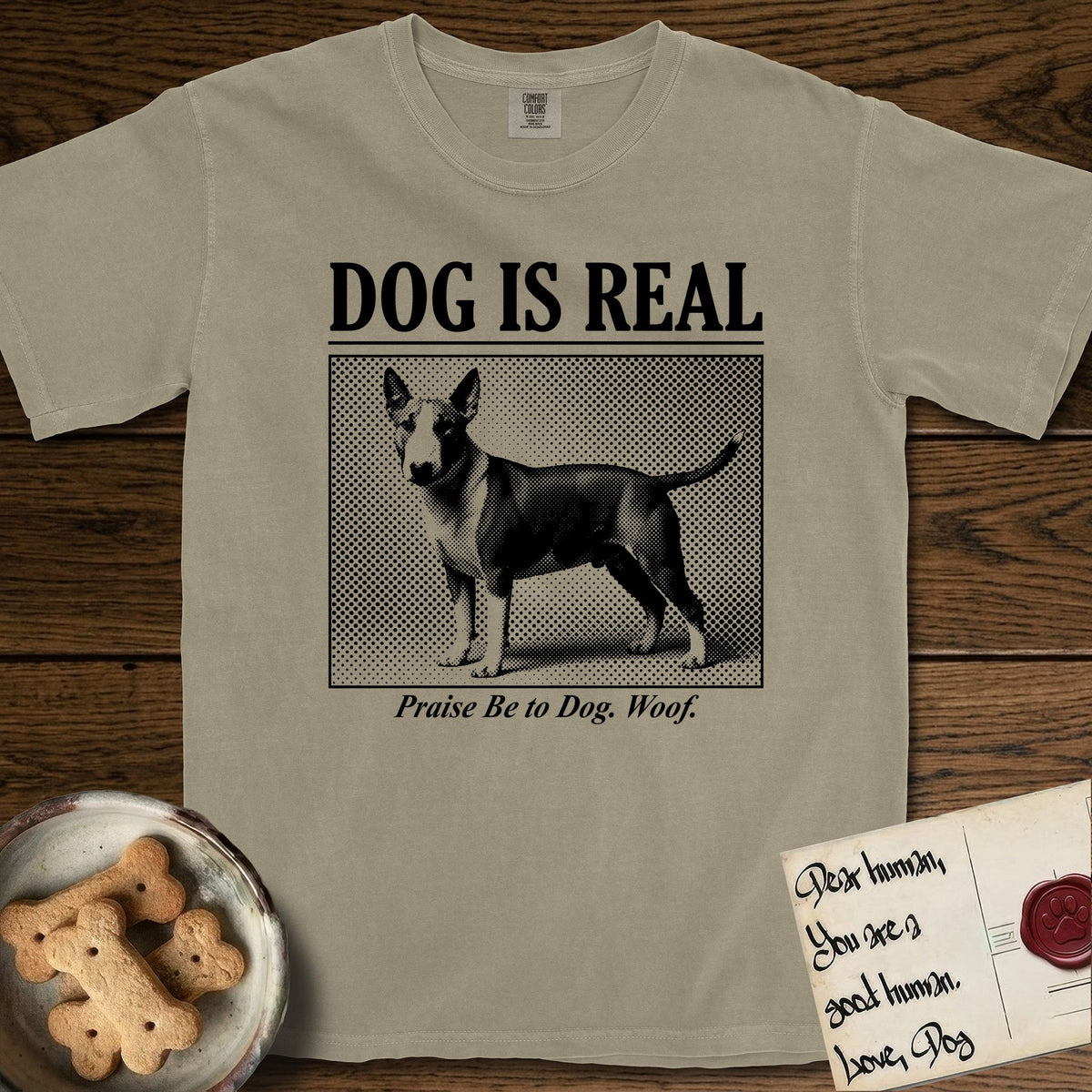 Dog is Real Bull Terrier T-Shirt flat lay in Khaki, featuring Black halftone newspaper photo of a Bull Terrier with a white face standing in profile with head turned to the camera, under the headline DOG IS REAL, with the caption Praise Be to Dog. Woof..