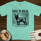 Dog is Real Cairn Terrier T-Shirt flat lay in Island Reef, featuring Black halftone newspaper photo of a shaggy Cairn Terrier with upright ears and tail standing in profile with head turned to the camera, under the headline DOG IS REAL, with the caption Praise Be to Dog. Woof..