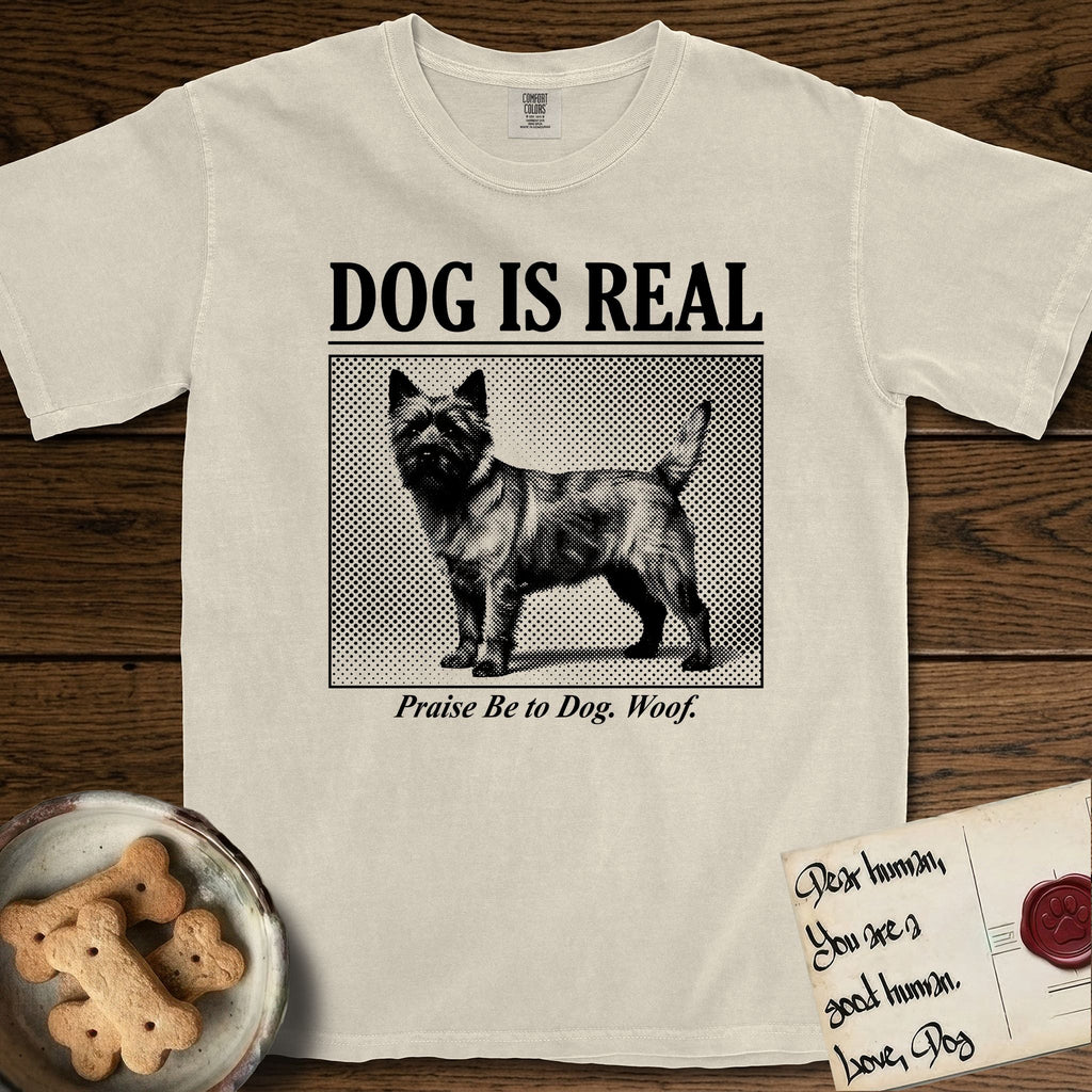 Dog is Real Cairn Terrier T-Shirt flat lay in Ivory, featuring Black halftone newspaper photo of a shaggy Cairn Terrier with upright ears and tail standing in profile with head turned to the camera, under the headline DOG IS REAL, with the caption Praise Be to Dog. Woof..