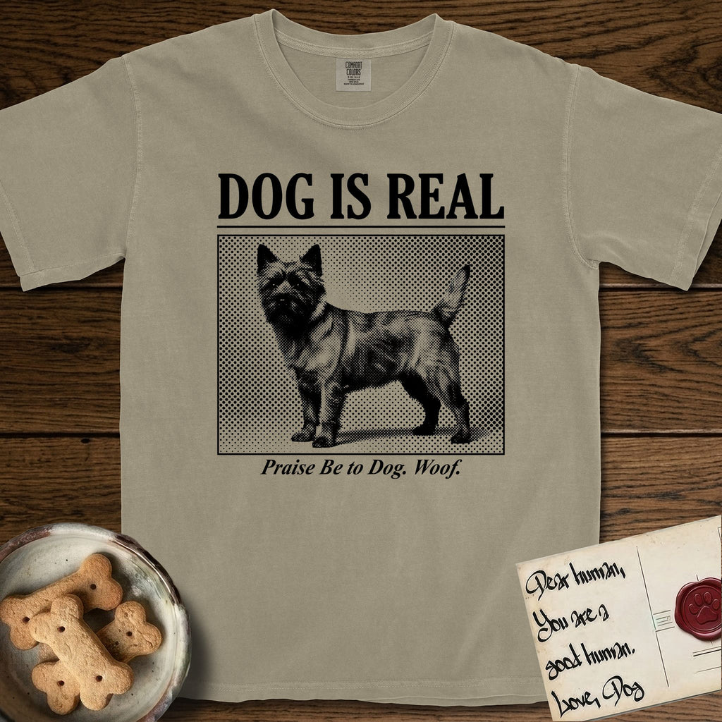 Dog is Real Cairn Terrier T-Shirt flat lay in Khaki, featuring Black halftone newspaper photo of a shaggy Cairn Terrier with upright ears and tail standing in profile with head turned to the camera, under the headline DOG IS REAL, with the caption Praise Be to Dog. Woof..