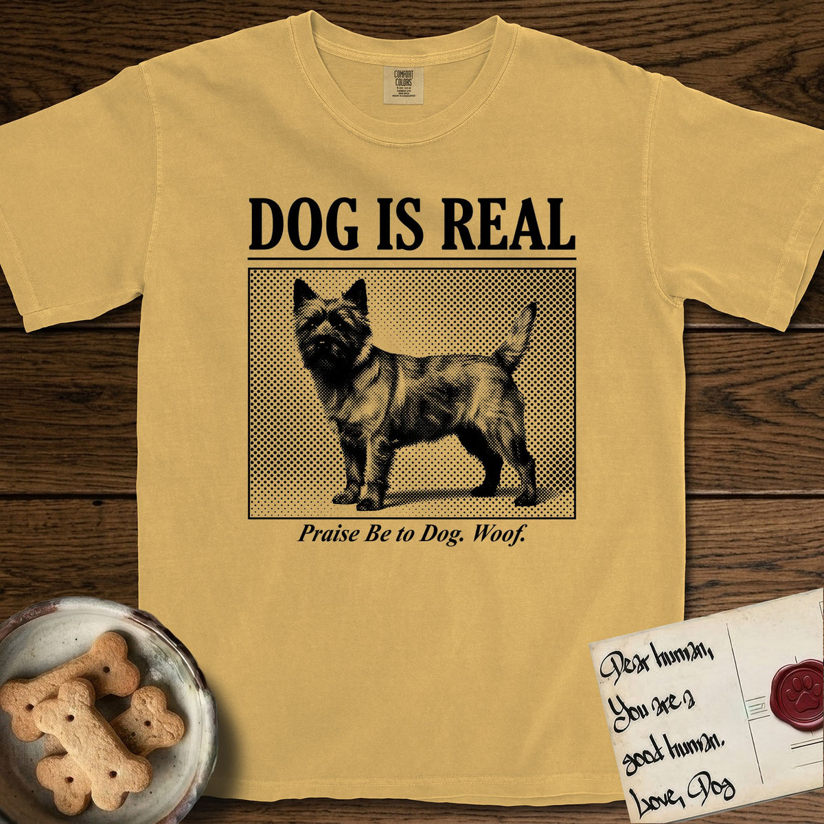 Dog is Real Cairn Terrier T-Shirt flat lay in Mustard, featuring Black halftone newspaper photo of a shaggy Cairn Terrier with upright ears and tail standing in profile with head turned to the camera, under the headline DOG IS REAL, with the caption Praise Be to Dog. Woof..