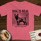 Dog is Real Cairn Terrier T-Shirt flat lay in Watermelon, featuring Black halftone newspaper photo of a shaggy Cairn Terrier with upright ears and tail standing in profile with head turned to the camera, under the headline DOG IS REAL, with the caption Praise Be to Dog. Woof..