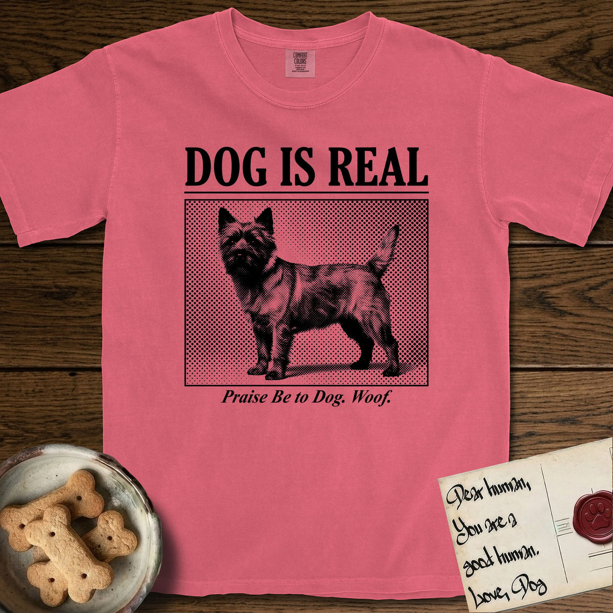 Dog is Real Cairn Terrier T-Shirt flat lay in Watermelon, featuring Black halftone newspaper photo of a shaggy Cairn Terrier with upright ears and tail standing in profile with head turned to the camera, under the headline DOG IS REAL, with the caption Praise Be to Dog. Woof..