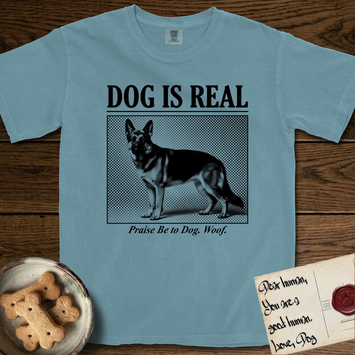 Dog is Real German Shepherd T-Shirt flat lay in Ice Blue, featuring Black halftone newspaper photo of a black and tan German Shepherd with upright ears standing in profile with head turned to the camera, under the headline DOG IS REAL, with the caption Praise Be to Dog. Woof..