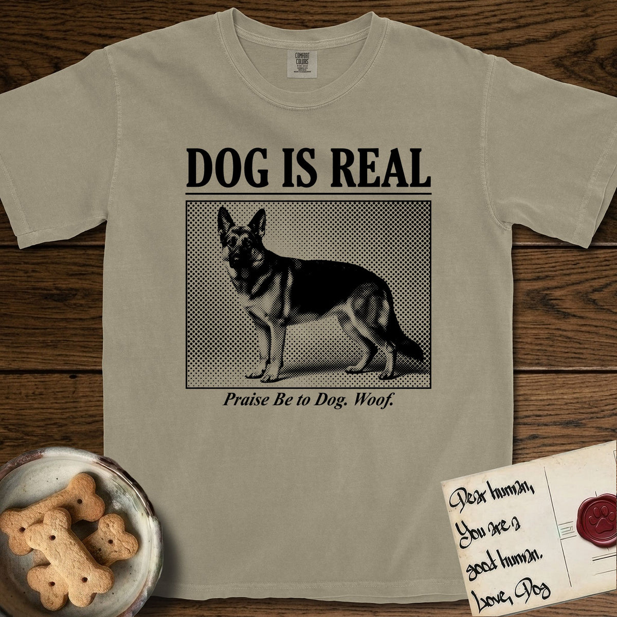 Dog is Real German Shepherd T-Shirt flat lay in Khaki, featuring Black halftone newspaper photo of a black and tan German Shepherd with upright ears standing in profile with head turned to the camera, under the headline DOG IS REAL, with the caption Praise Be to Dog. Woof..