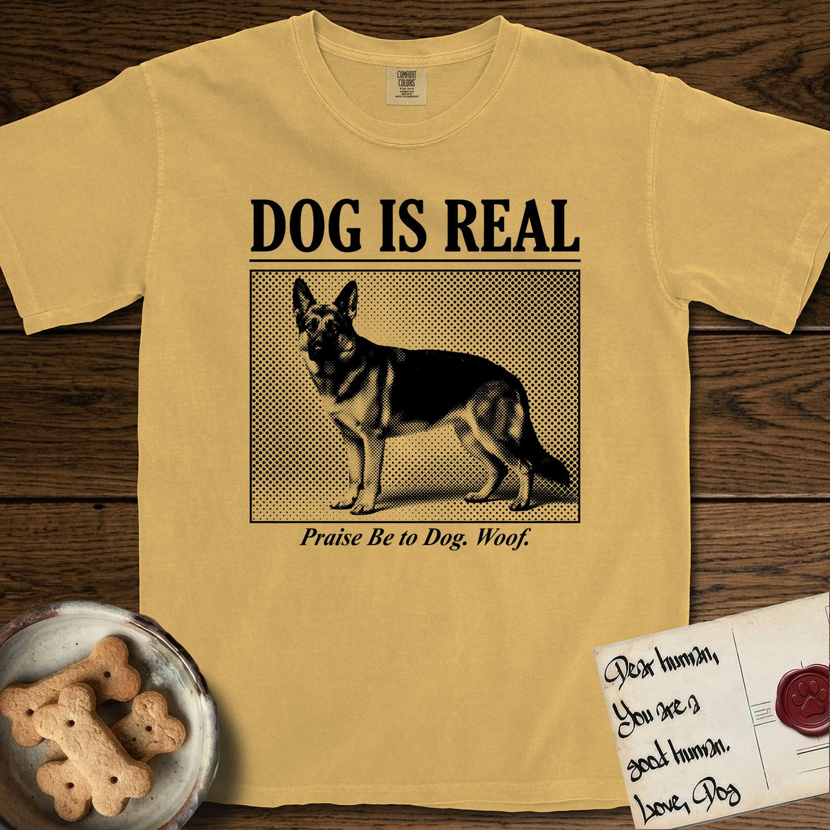 Dog is Real German Shepherd T-Shirt flat lay in Mustard, featuring Black halftone newspaper photo of a black and tan German Shepherd with upright ears standing in profile with head turned to the camera, under the headline DOG IS REAL, with the caption Praise Be to Dog. Woof..