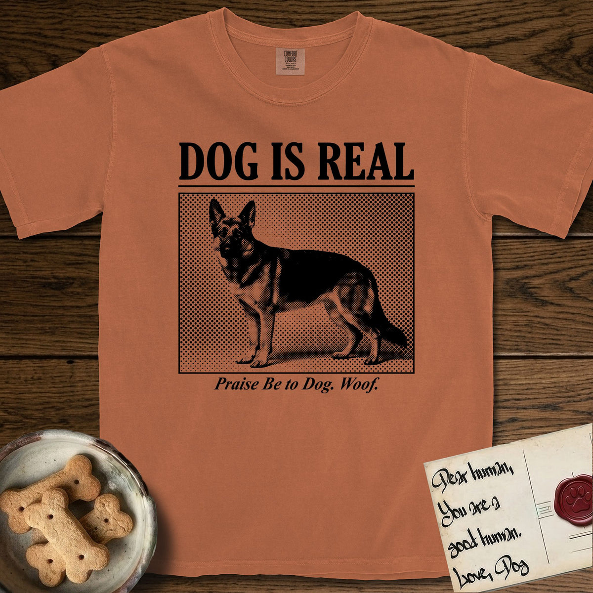 Dog is Real German Shepherd T-Shirt flat lay in Yam, featuring Black halftone newspaper photo of a black and tan German Shepherd with upright ears standing in profile with head turned to the camera, under the headline DOG IS REAL, with the caption Praise Be to Dog. Woof..
