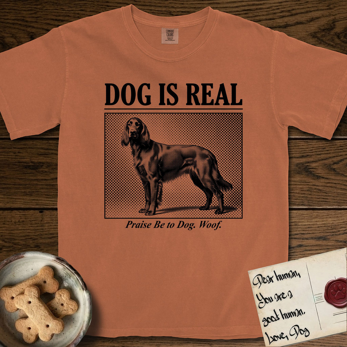 Dog is Real Irish Setter T-Shirt flat lay in Yam, featuring Black halftone newspaper photo of a feathered Irish Setter standing in profile with head turned to the camera, under the headline DOG IS REAL, with the caption Praise Be to Dog. Woof..