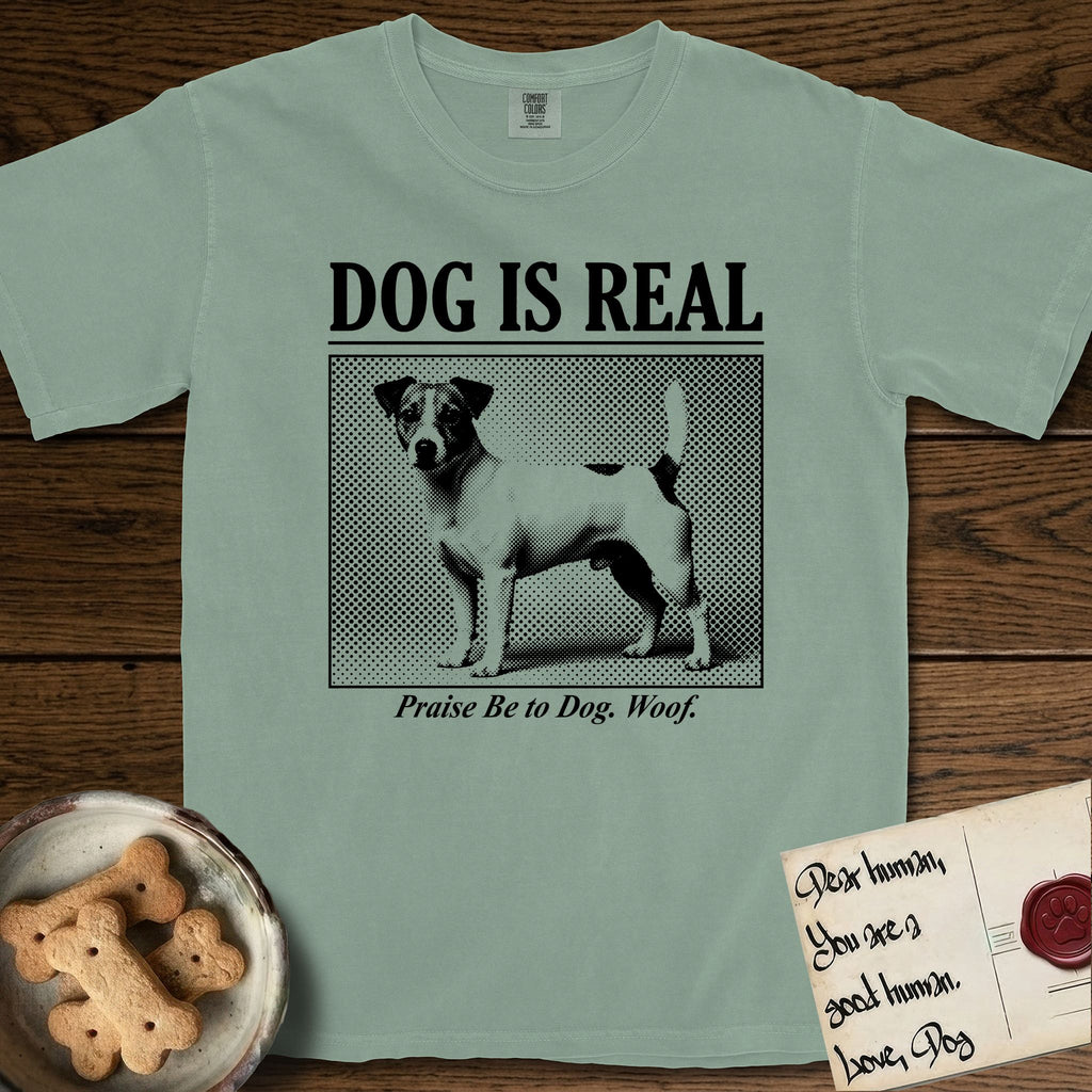 Dog is Real Jack Russell T-Shirt flat lay in Bay, featuring Black halftone newspaper photo of a mostly white Jack Russell with dark ears and an upright tail standing in profile with head turned to the camera, under the headline DOG IS REAL, with the caption Praise Be to Dog. Woof..