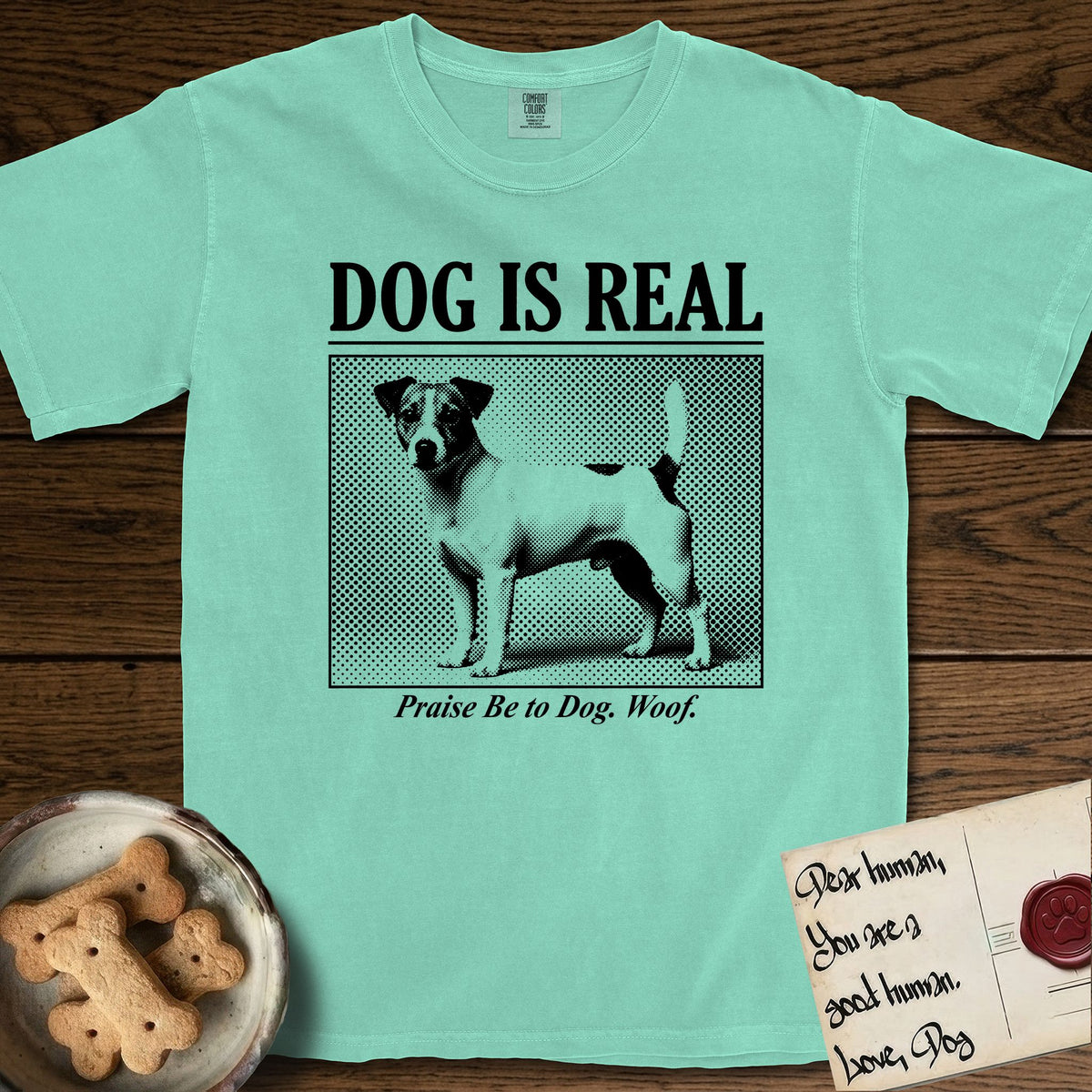Dog is Real Jack Russell T-Shirt flat lay in Island Reef, featuring Black halftone newspaper photo of a mostly white Jack Russell with dark ears and an upright tail standing in profile with head turned to the camera, under the headline DOG IS REAL, with the caption Praise Be to Dog. Woof..